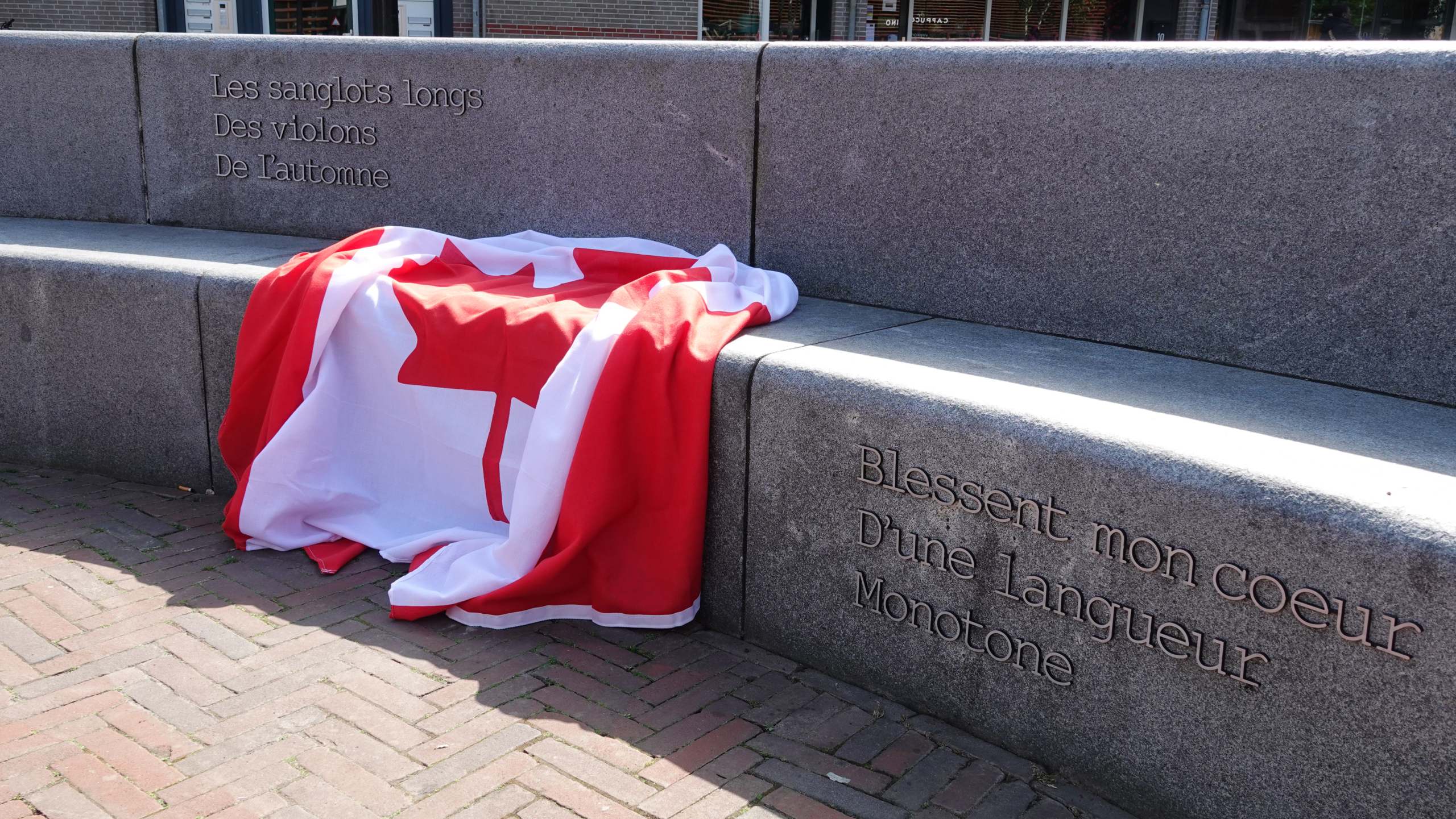 Een grijze stenen bank met een citaat in het Frans gegraveerd, bedekt met een Canadese vlag, op een plek met bakstenen bestrating.