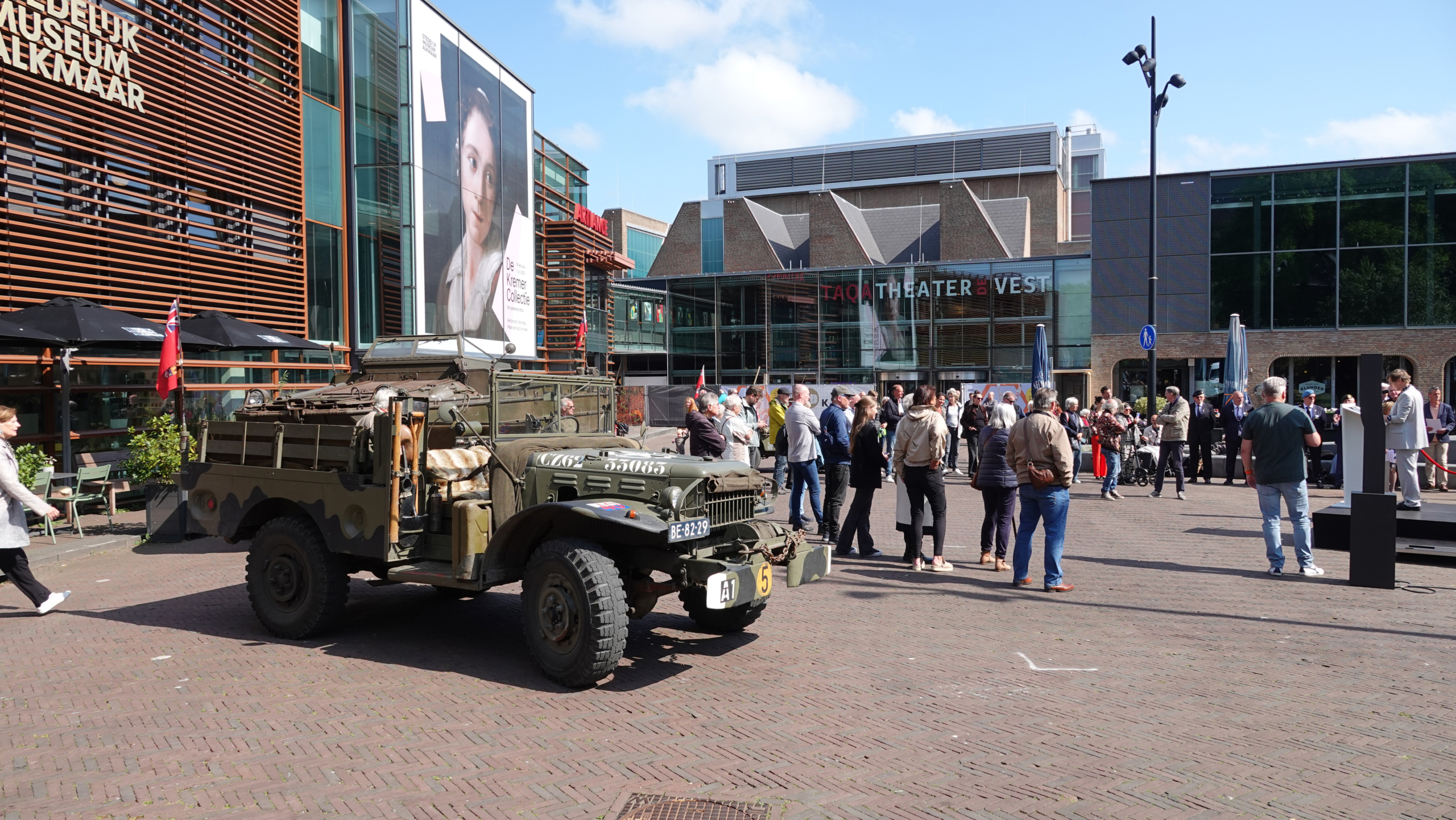 Een oude militaire jeep staat geparkeerd voor het Stedelijk Museum Alkmaar, waar mensen buiten verzamelen voor een evenement, met het TAQA Theater De Vest op de achtergrond.