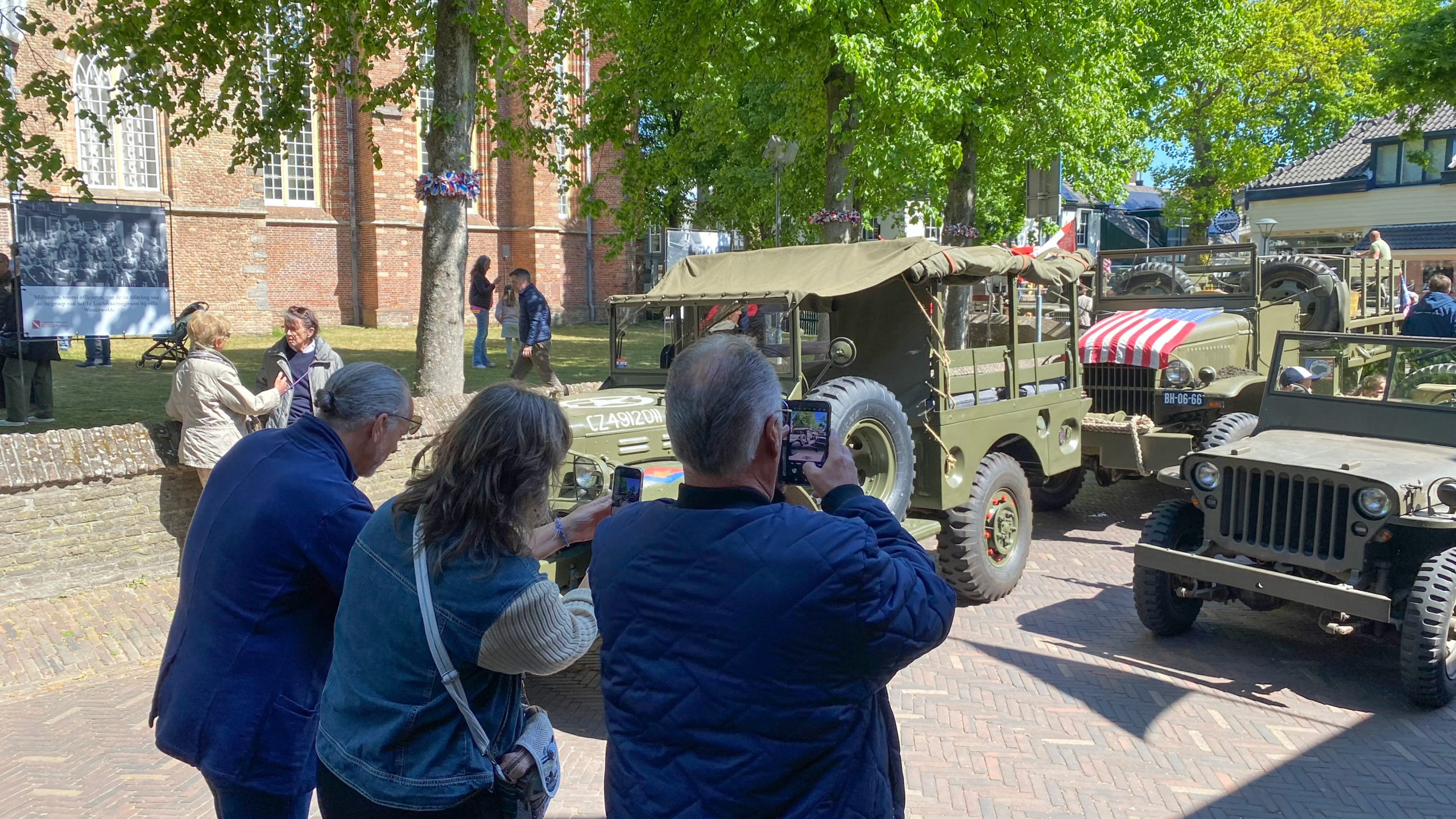Mensen maken foto's van oude militaire voertuigen die geparkeerd staan langs een straat, met een historisch gebouw op de achtergrond en bomen die schaduw bieden.