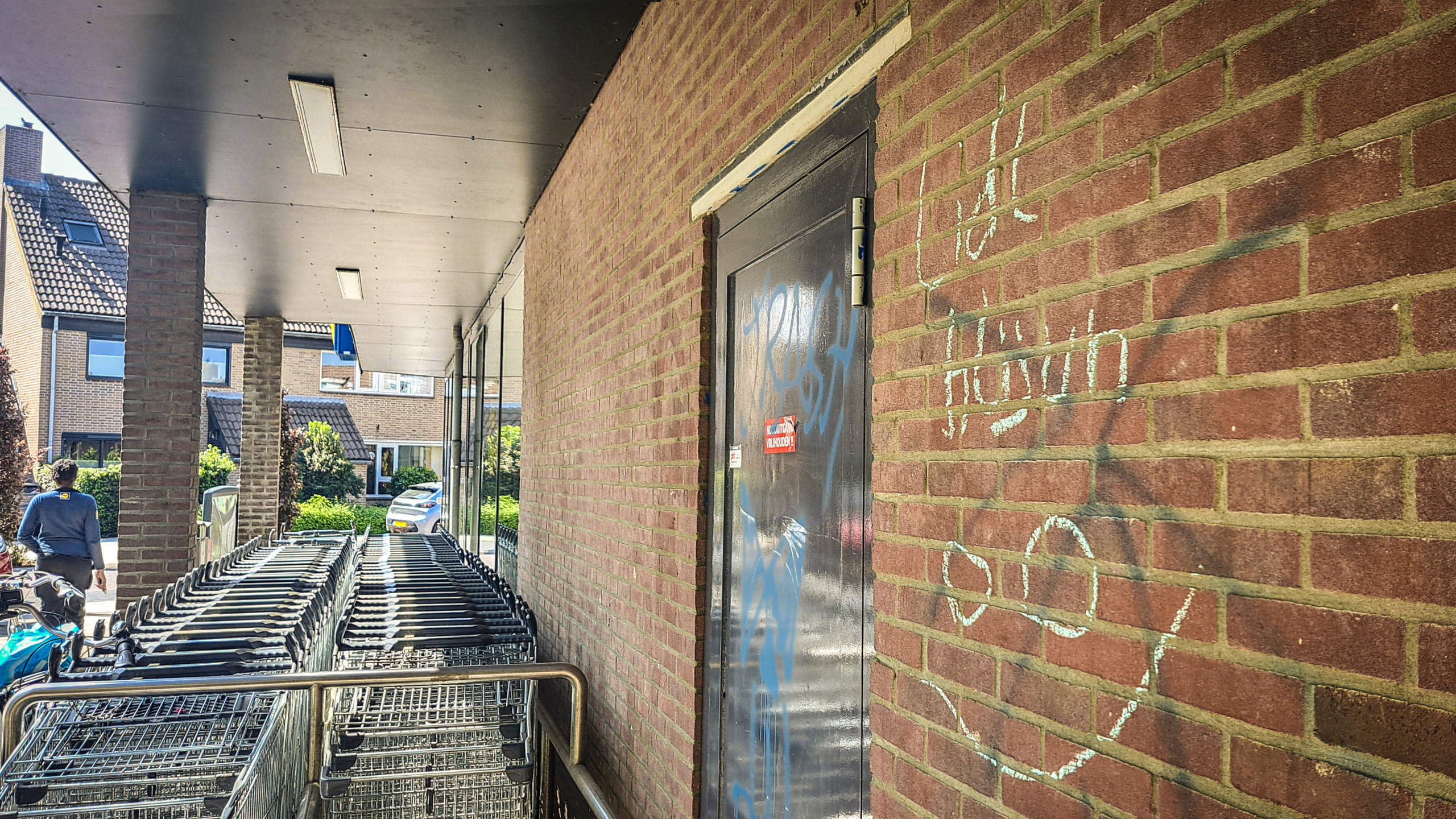 Een rij winkelwagens naast een bakstenen muur met graffiti, bij een overdekte ingang van een gebouw. Een persoon loopt weg van de muur, in de richting van de winkelwagens.