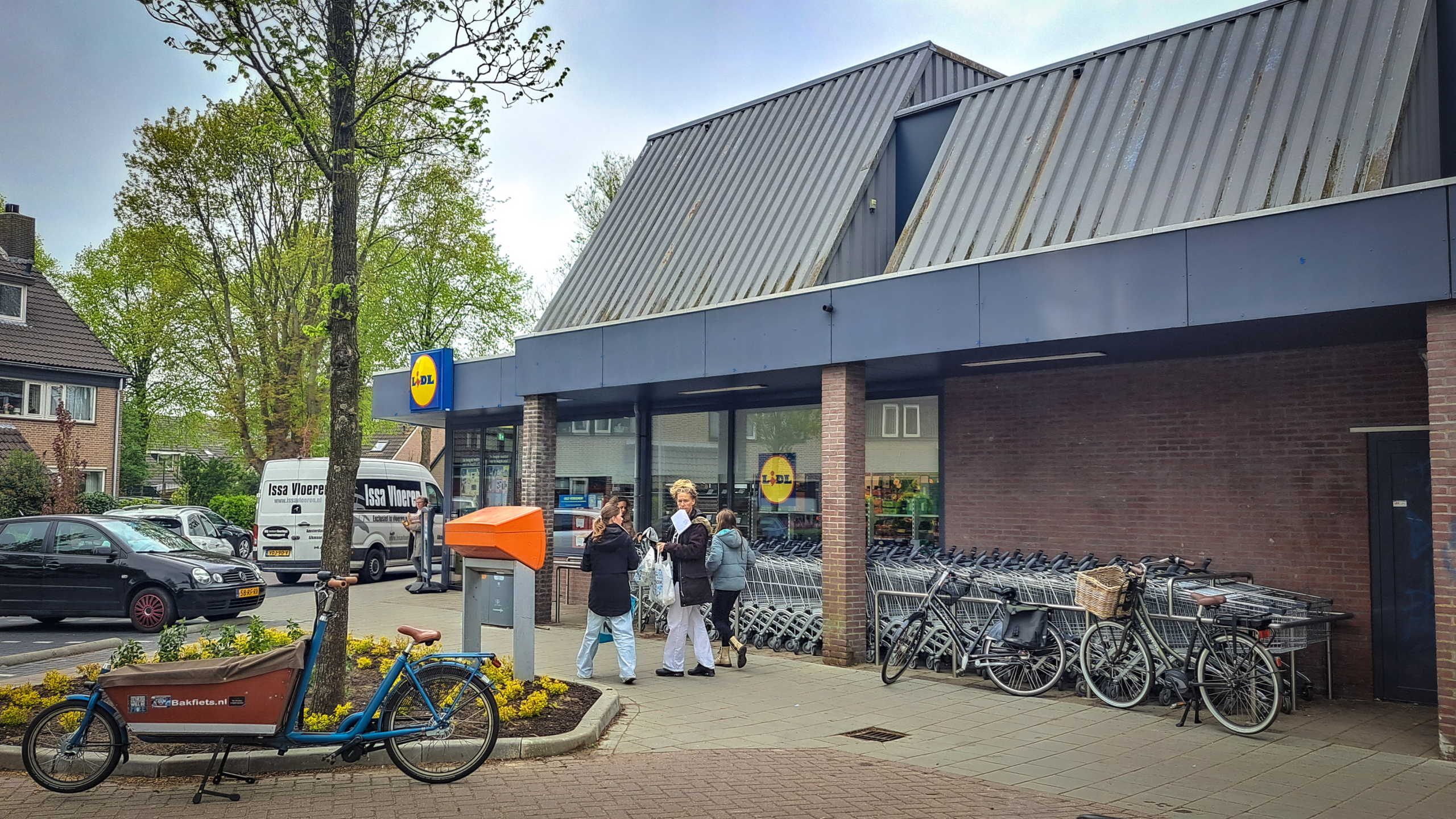 Een supermarkt met het logo van Lidl, enkele mensen staan bij winkelwagens bij de ingang, er zijn fietsen en een bakfiets geparkeerd, en een bestelbus in de achtergrond.