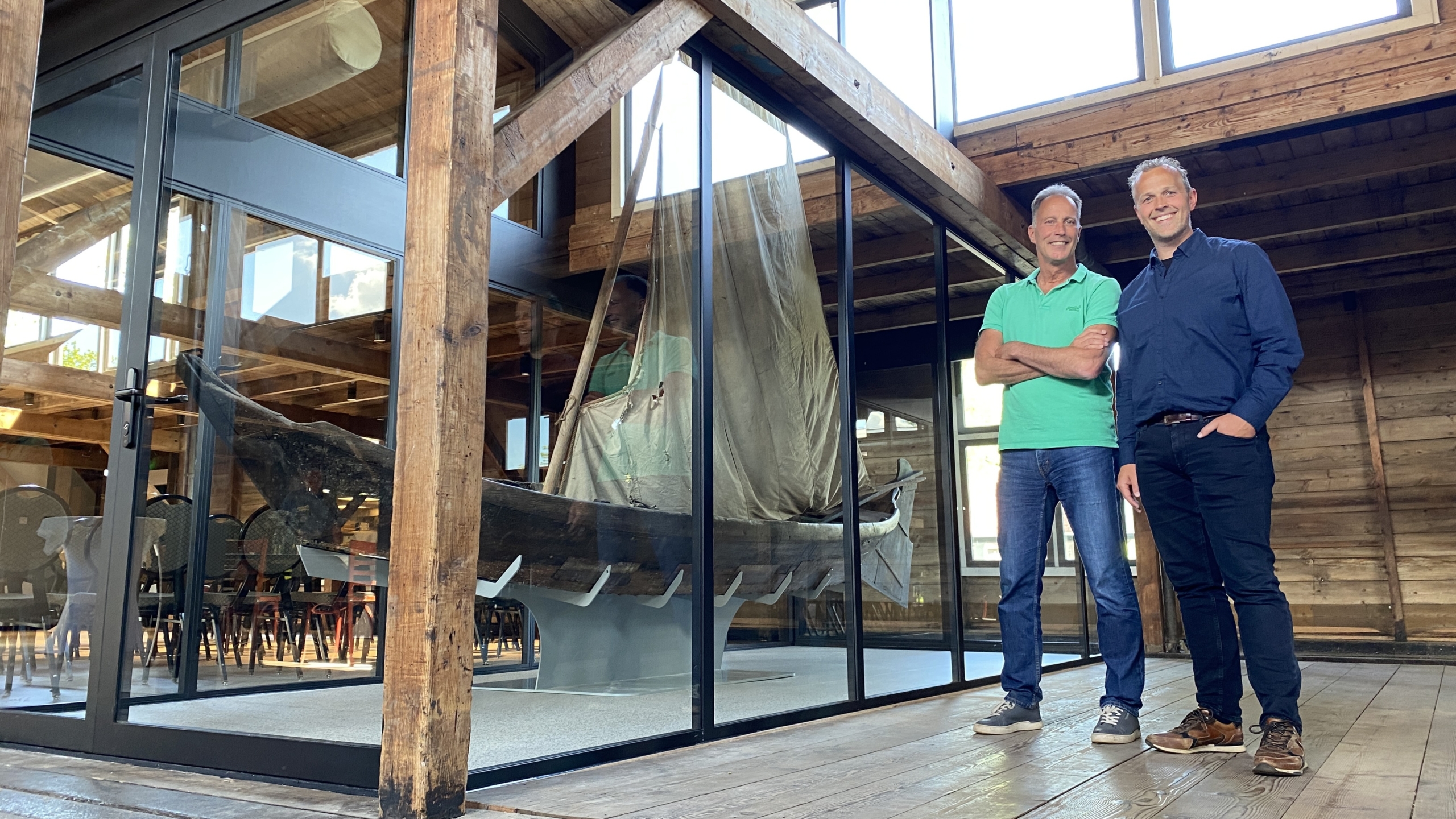 Twee mannen staan naast elkaar in een houten gebouw met een glazen wand, waarachter een traditionele houten boot met een opgetuigd zeil te zien is.