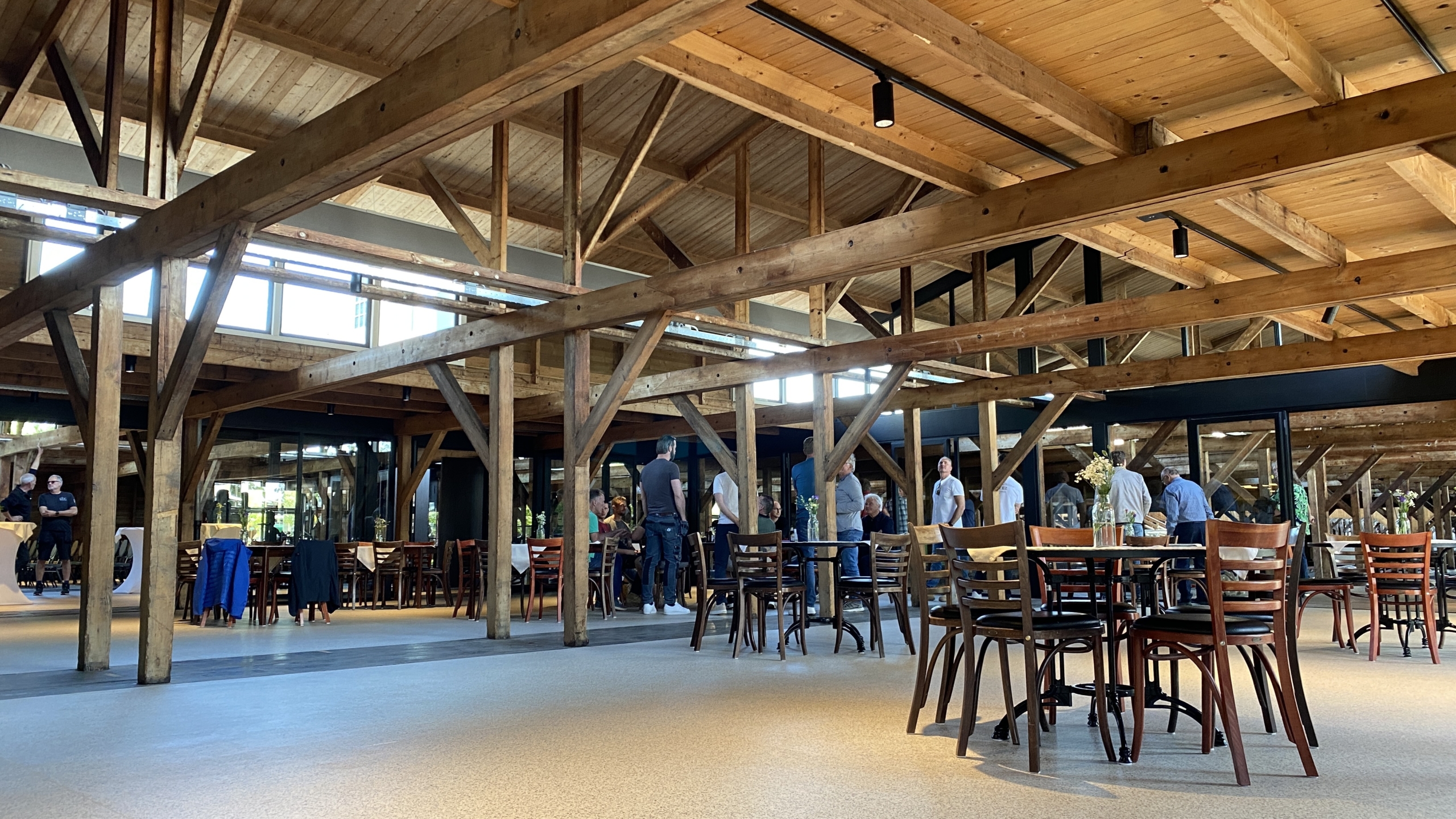 Interieur van een ruime houten zaal met zichtbare balken en houten tafels en stoelen; enkele mensen staan in de verte te praten.