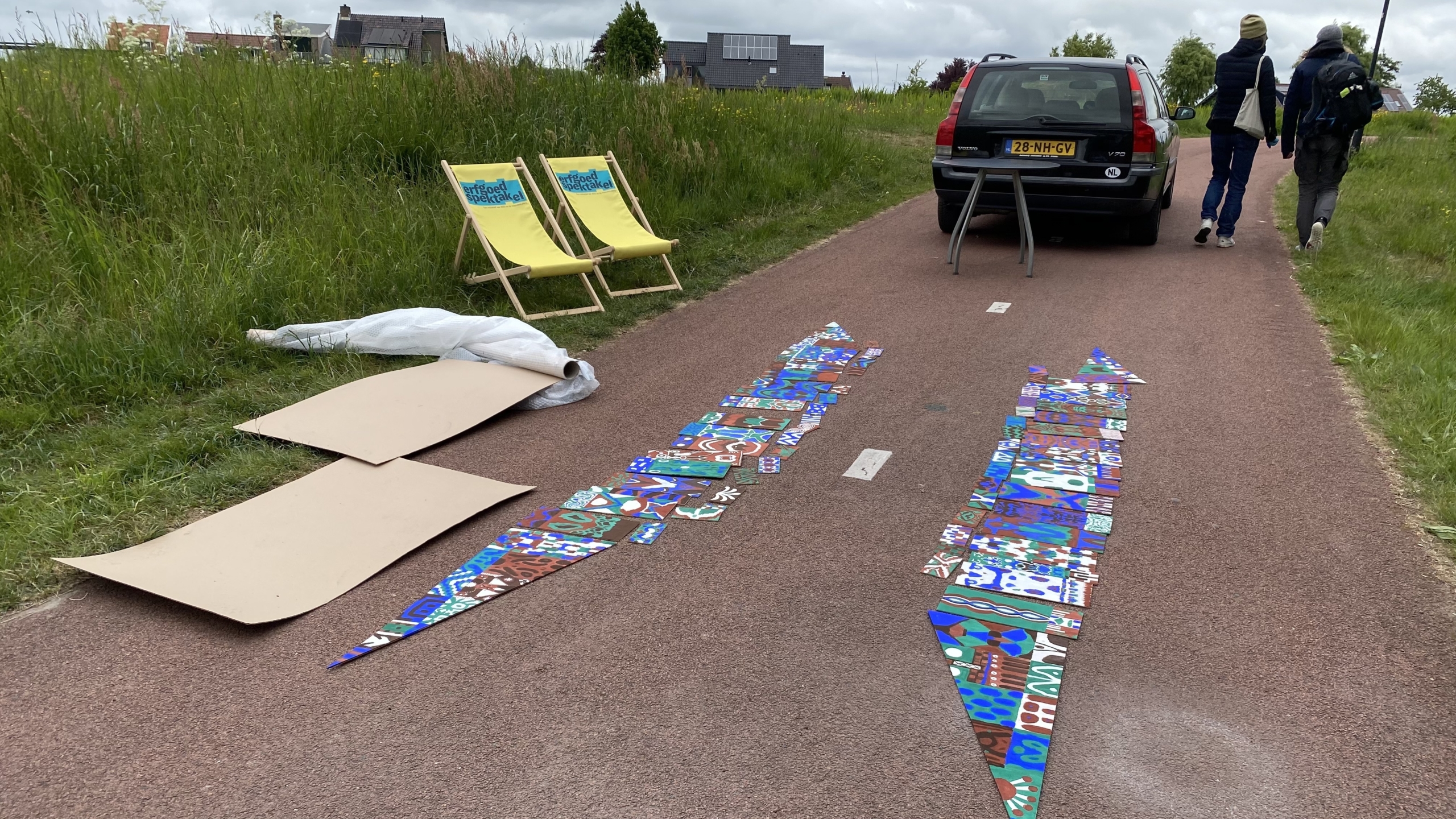 Twee gele strandstoelen en kartonnen stukken liggen op een fietspad naast een rode auto. Op het pad liggen kleurrijke, met patronen beschilderde stukken papier of stof in de vorm van pijlen. Twee personen lopen verderop naast de auto.