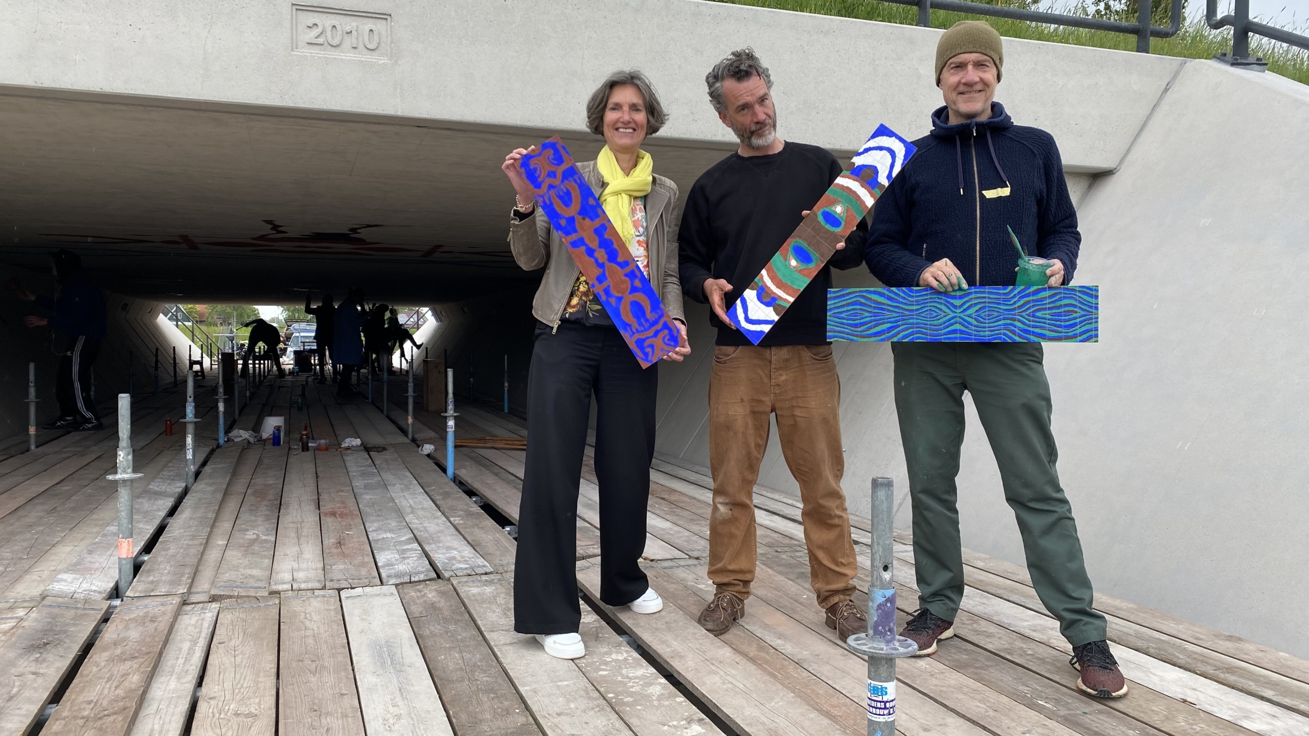 Drie mensen staan voor een onderdoorgang terwijl ze beschilderde houten panelen vasthouden; achter hen is een verhoogde houten plankier zichtbaar.