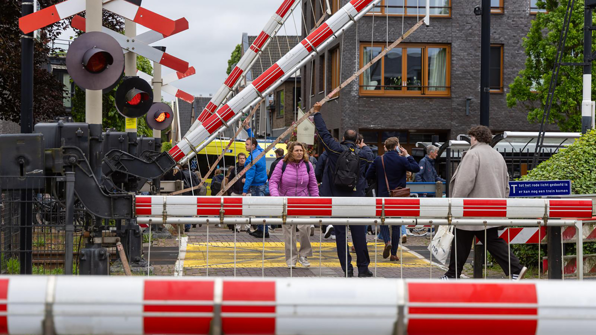 Mensen lopen over een overweg terwijl de slagbomen omhoog zijn en de waarschuwingslichten branden, met gebouwen op de achtergrond.