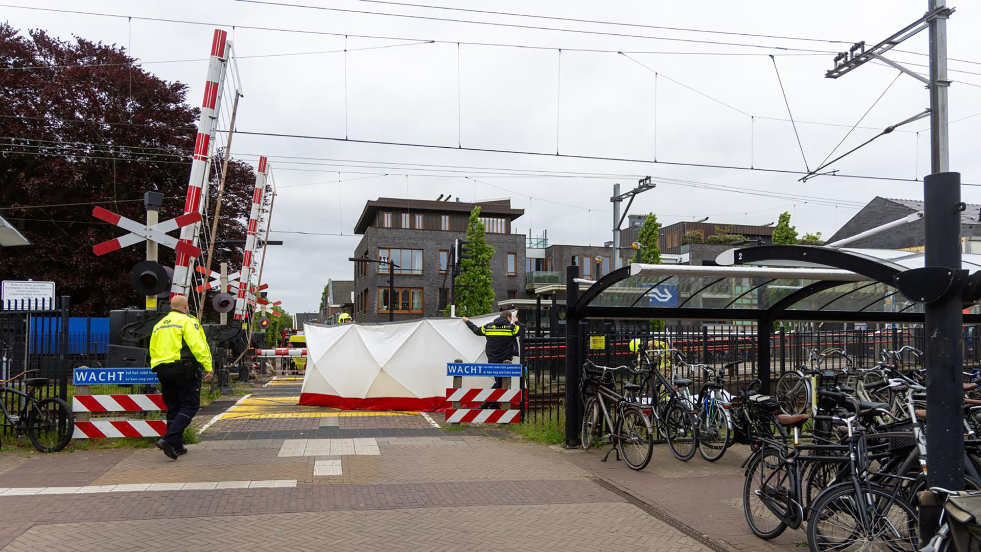 Treinoverweg met slagbomen gesloten en een witte tent geplaatst, beveiligd door politieagenten in gele jassen; fietsenrek aan de rechterkant en moderne gebouwen op de achtergrond.