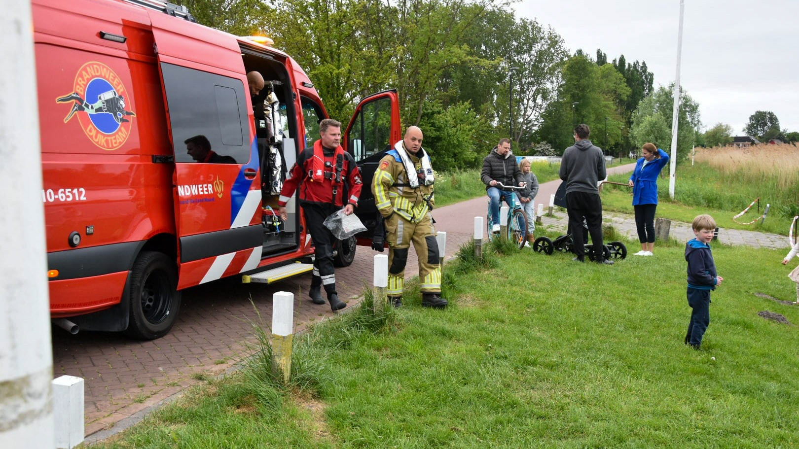 Een brandweerwagen staat geparkeerd naast een fietspad, met brandweermannen die uit het voertuig stappen, in een groene buitenomgeving met enkele mensen in de buurt.