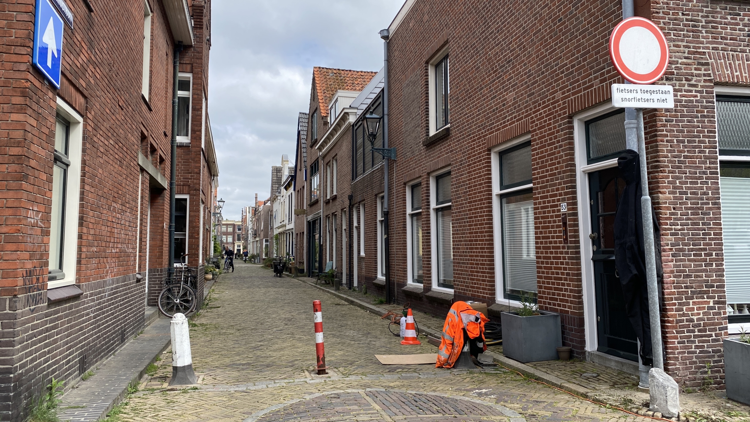 Een smalle straat met rode bakstenen huizen aan weerszijden, een verkeersbord met de tekst "fietsers toegestaan, snorfietsers niet," enkele verkeerskegels en een fiets geparkeerd tegen een muur.
