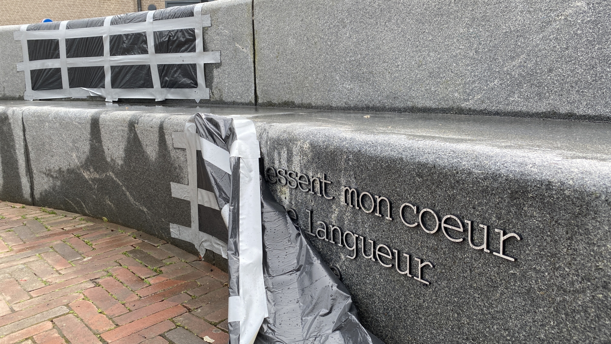 Een stenen monument met Franse tekst erop, gedeeltelijk bedekt met zwarte plastic zakken die met ducttape zijn vastgezet. Het bevindt zich naast een bakstenen trottoir.