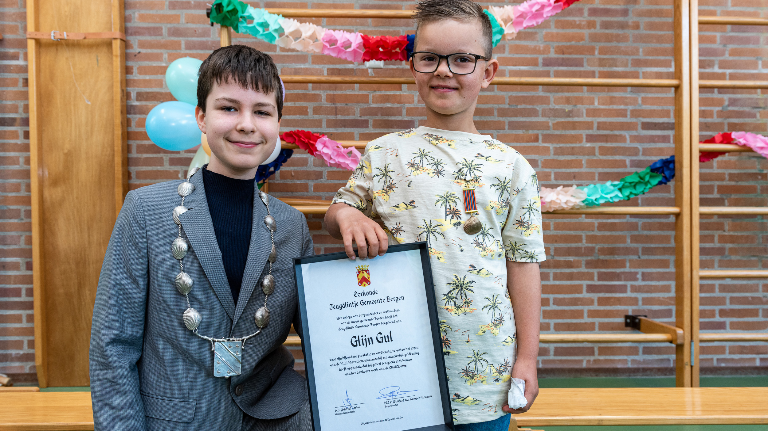 Twee jongens poseren samen; de linker draagt een pak met ketting en de rechter houdt een oorkonde vast en draagt een t-shirt met palmbomen. Achter hen hangen gekleurde slingers en ballonnen tegen een bakstenen muur.