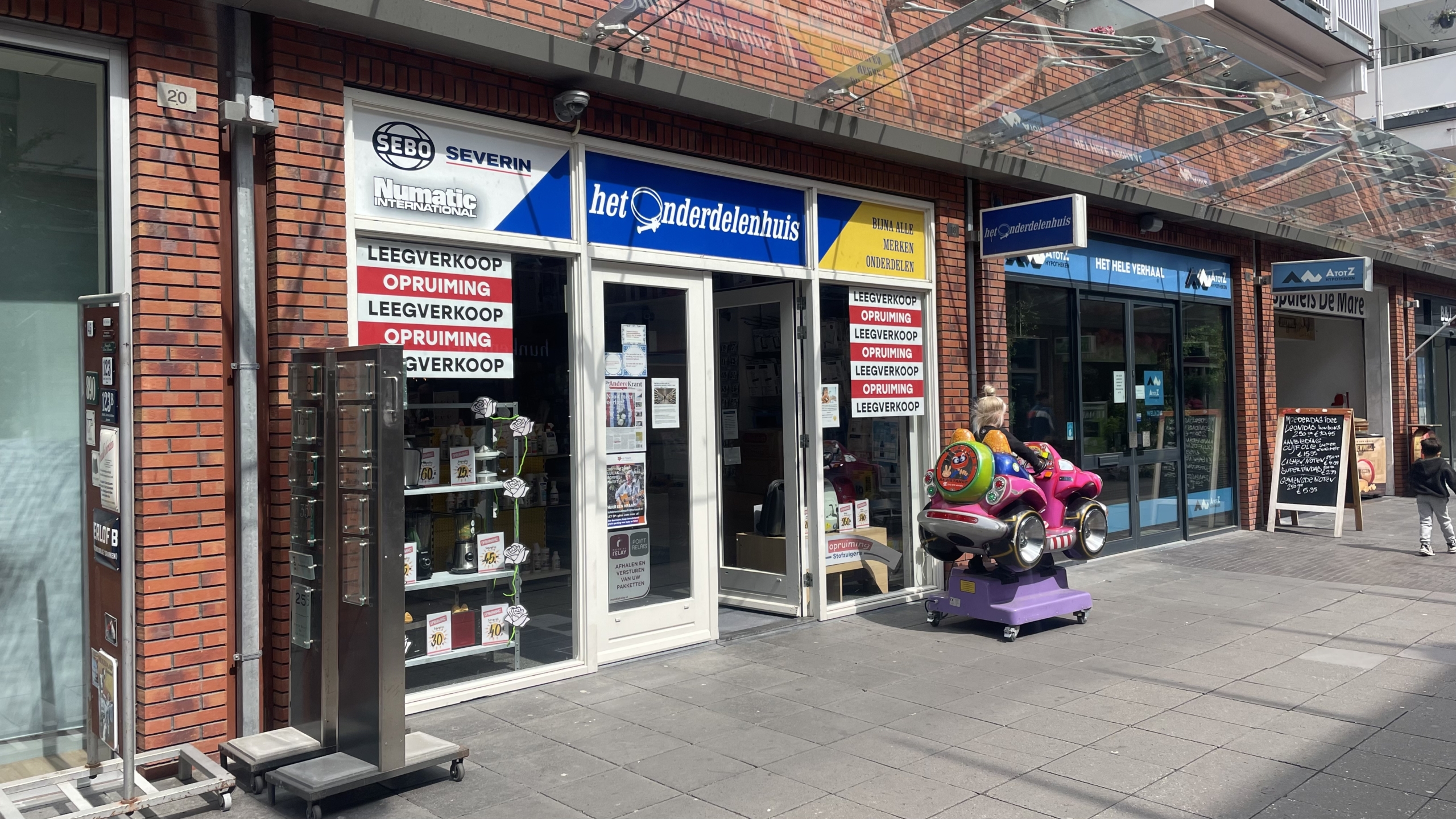 Straatbeeld met een winkel met een bord "het onderdelenhuis" en een kind in een roze speeltuig voor de winkel. Er zijn raamstickers met "leegverkoop opruiming" en een glazen overkapping boven de ingang.