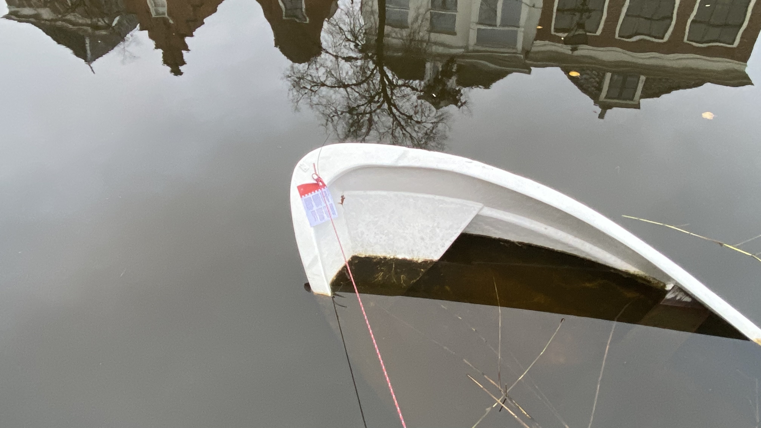 Een half gezonken wit bootje in een rustige gracht, met op de achtergrond de reflectie van huizen en bomen in het water.