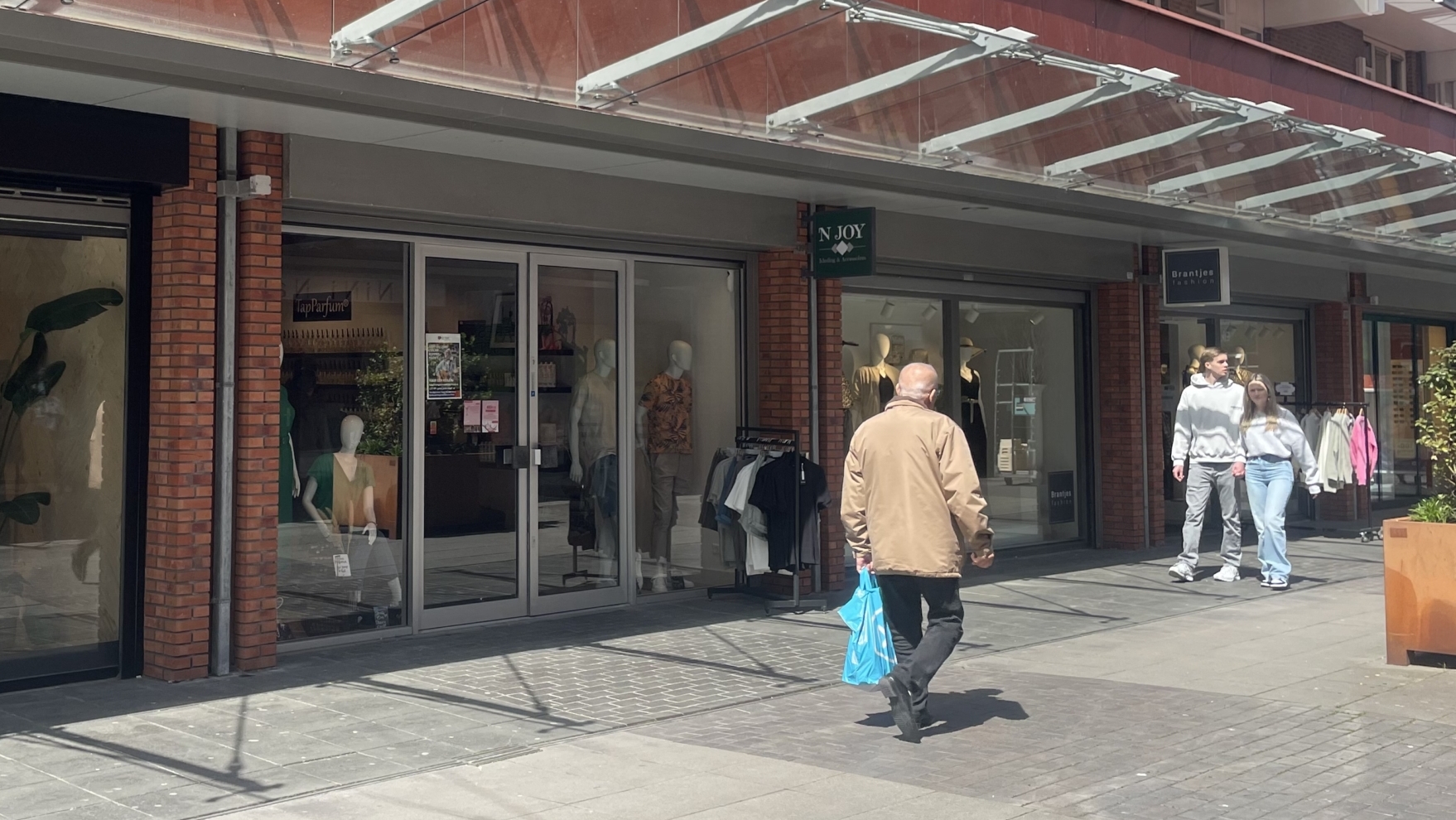 Winkelstraat met winkels links en ramen met etalages; een man en een vrouw lopen aan de rechterkant en een andere persoon met een blauwe tas loopt van de camera af.