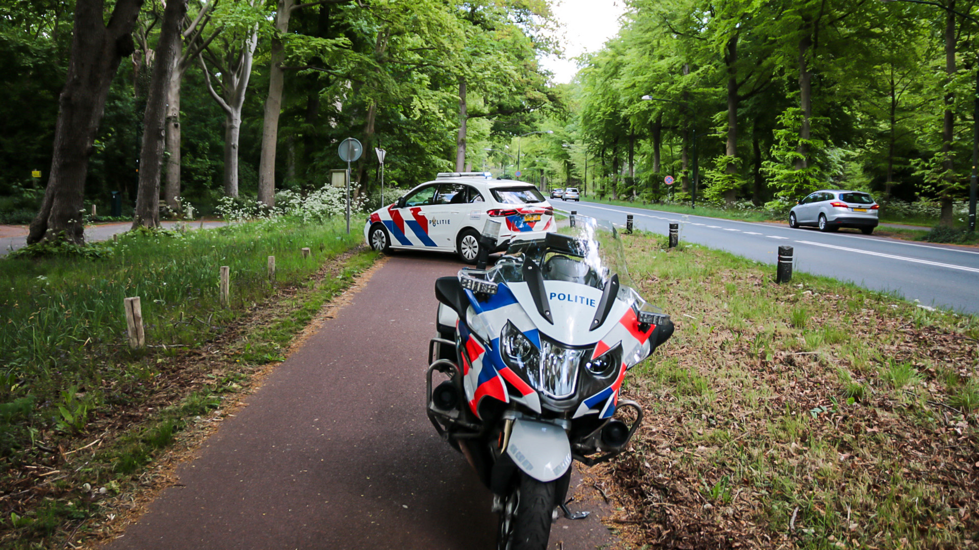 Een politieauto en politiemotor staan geparkeerd langs een bospad naast een landelijke weg, omgeven door bomen en groen gras.
