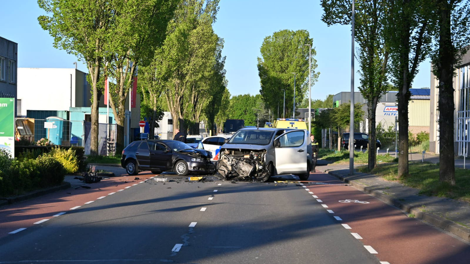 Twee auto's met schade aan de voorkant staan op een weg, een ervan heeft de motorkap open; er zijn brokstukken verspreid op het wegdek en een ambulance is aanwezig op de achtergrond.