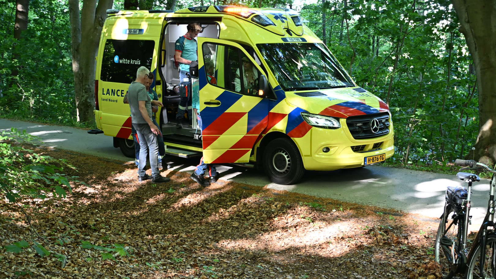 Gele ambulance van Witte Kruis met geopende deur op een bosweg, met een paramedicus en een man naast de ambulance en een fiets op de voorgrond.