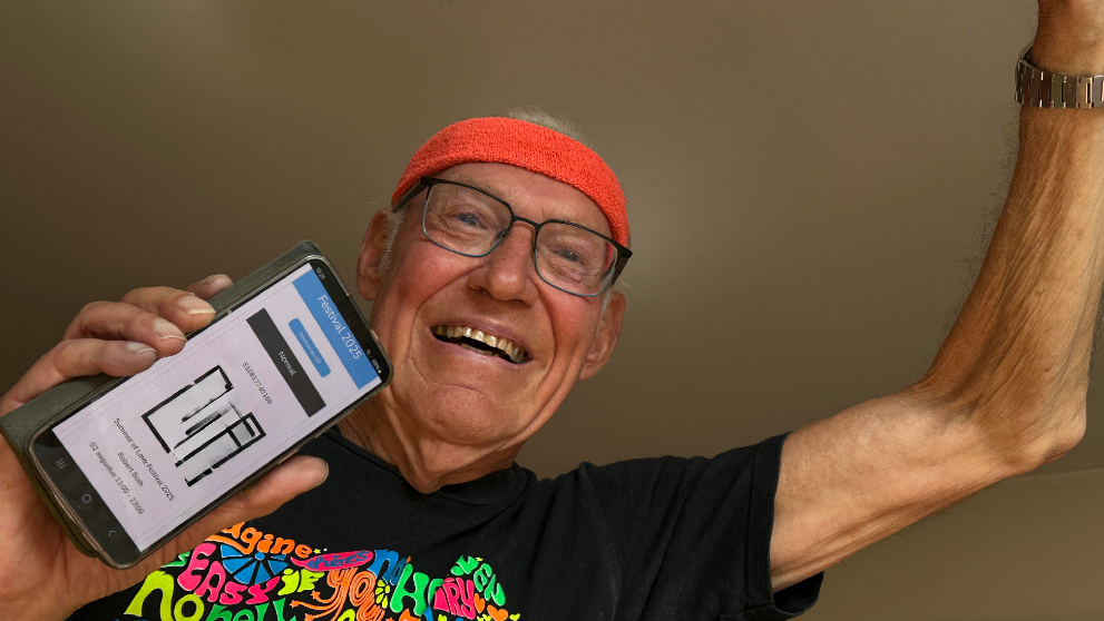 Oudere man met een rode hoofdband en een kleurrijk T-shirt houdt een smartphone omhoog waarop een festivalkaartje staat, met een glimlach op zijn gezicht.