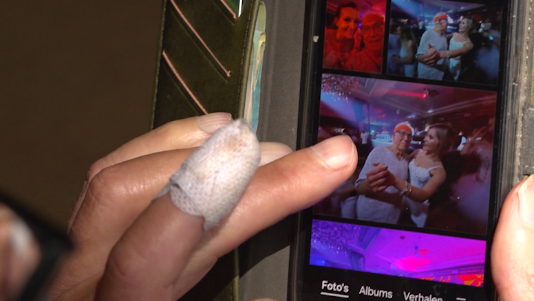 Een hand met een verband om een vinger navigeert door foto's op een smartphone. Het scherm toont verschillende afbeeldingen van mensen die samen poseren op een evenement. De woorden "Foto's", "Albums" en "Verhalen" zijn zichtbaar onderaan het scherm.