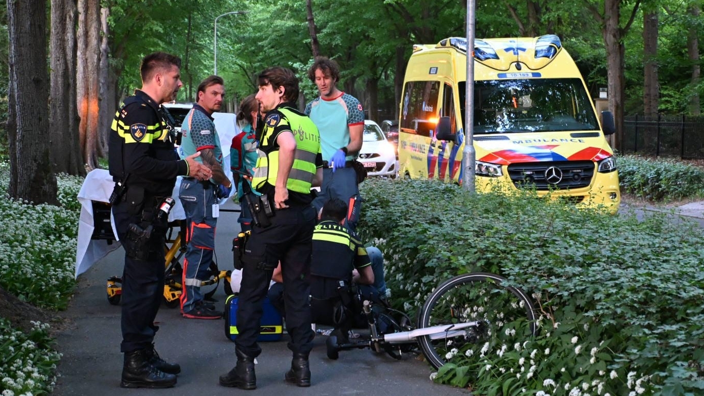 Politie en medisch personeel verlenen hulp bij een fietsongeval in een bosrijke omgeving, met een ambulance op de achtergrond.