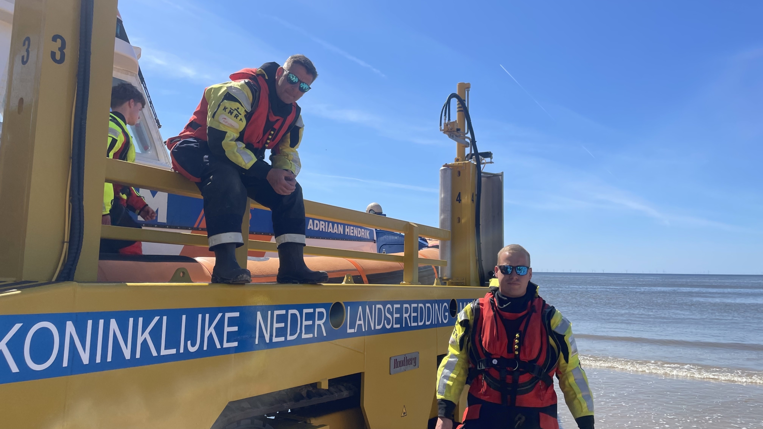 Twee leden van de KNRM in reddingskleding staan bij een reddingsvoertuig aan zee op een zonnige dag.