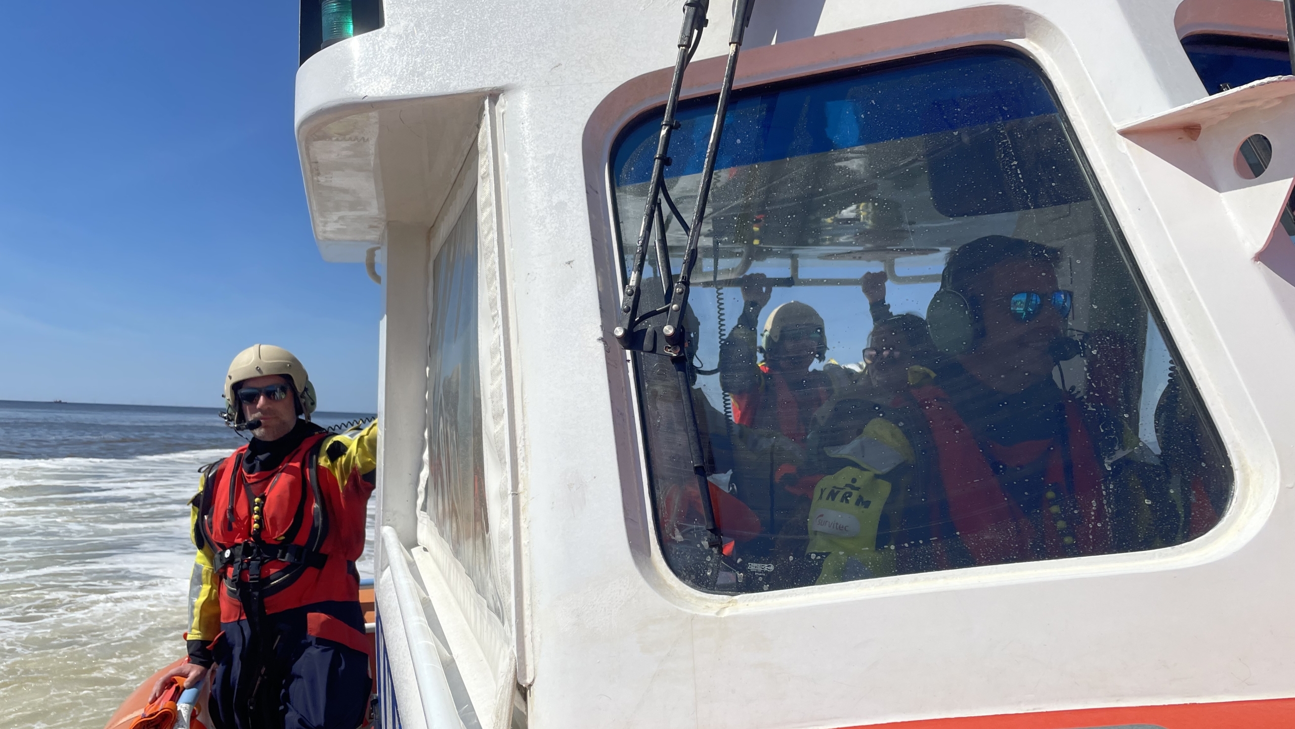Reddingswerkers in geel en oranje uitrusting aan boord van een reddingsboot op zee, met een man buiten staand tegen de cabine terwijl anderen binnen zitten, sommige met hun armen omhoog gejuich uitdrukkend.