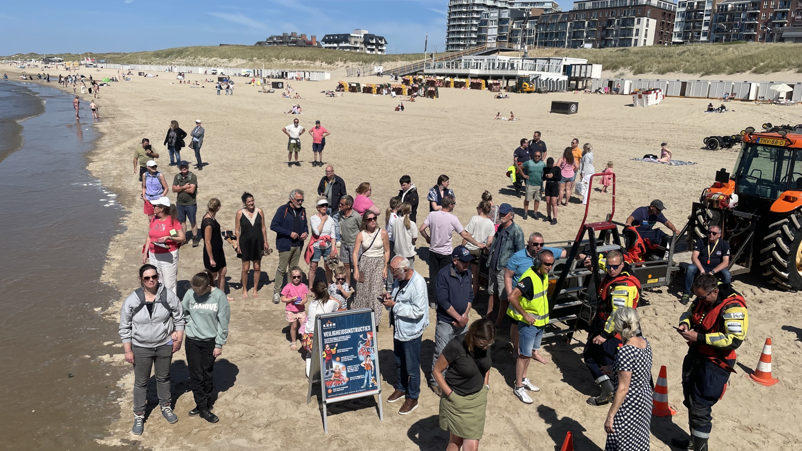 Grote groep mensen verzameld op een strand, waaronder strandgasten en personeel in reddingsvesten, naast een tractor en een bord met veiligheidsinstructies.