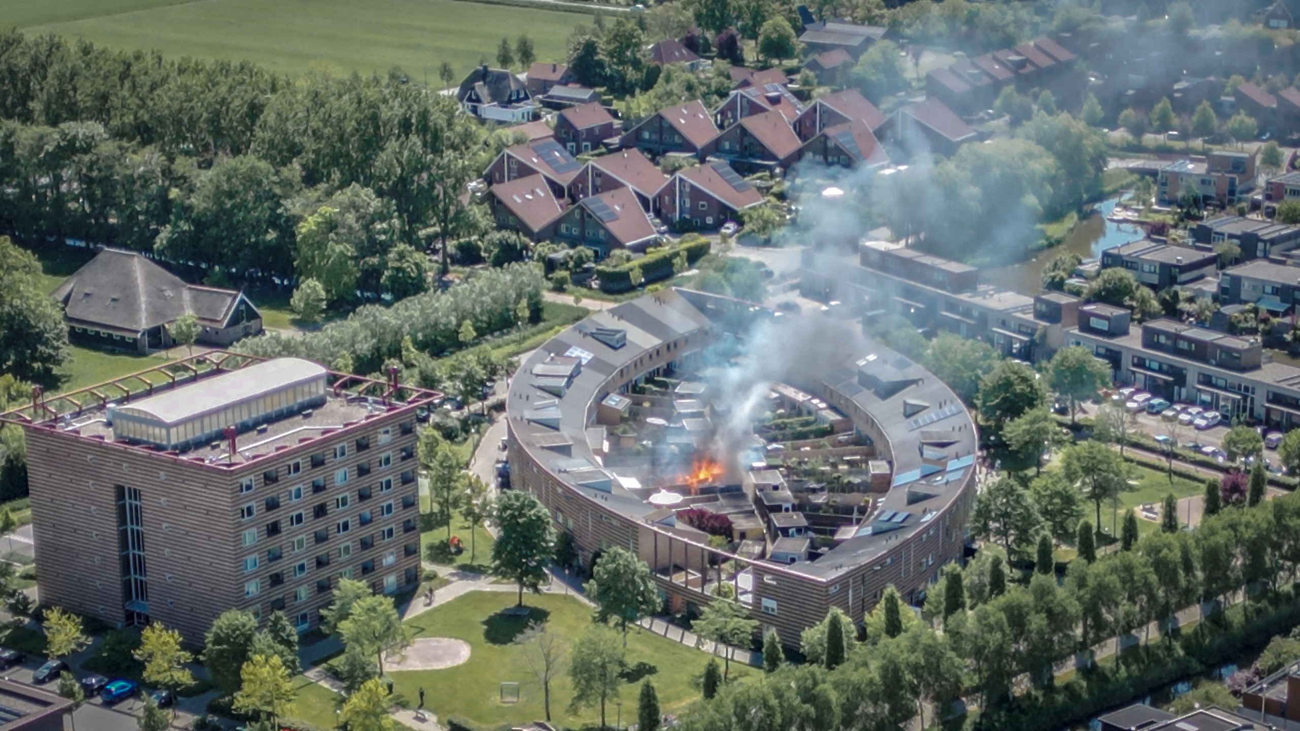 Luchtfoto van een gebogen appartementencomplex waar rook en vlammen zichtbaar zijn, omringd door een wijk met woningen en groen. Er is sprake van brand in het complex.