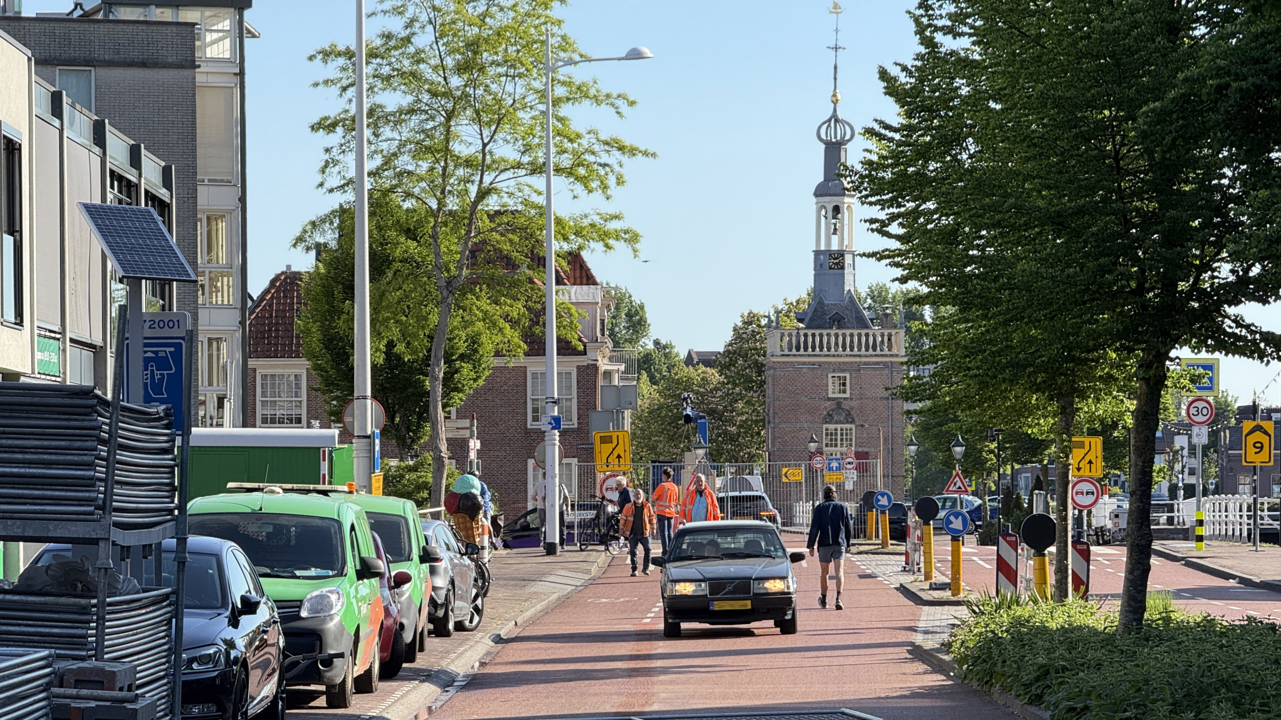 Straat in stedelijke omgeving met geparkeerde auto's, mensen die fietsen en lopen, en in de verte een historisch gebouw met een toren.