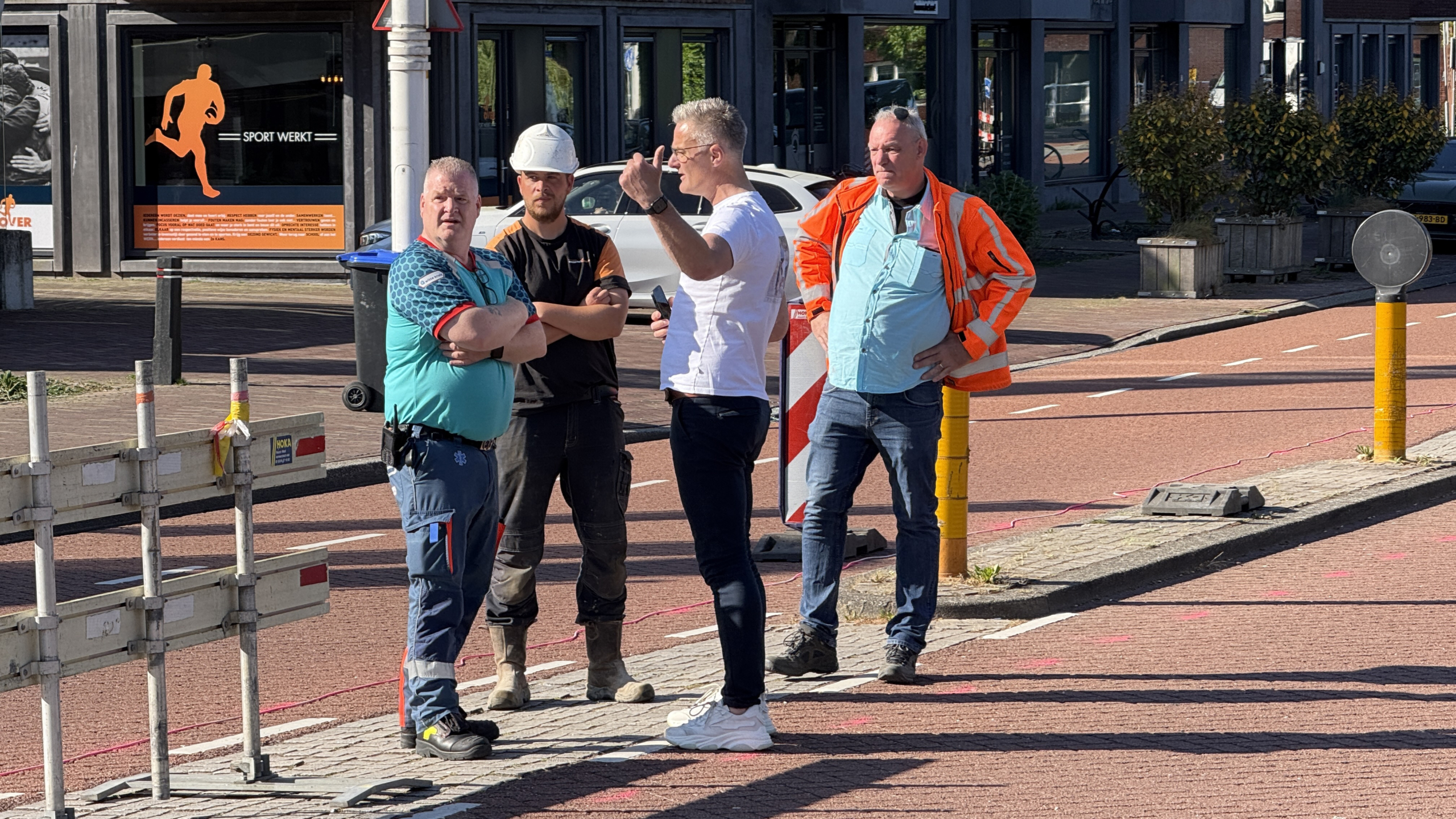 Vier mannen staan op een trottoir, waarvan een in een oranje veiligheidsjas, een in een bouwhelm, en een in een blauw T-shirt. Ze lijken een discussie of overleg te voeren naast een weg met verkeersbarrières.
