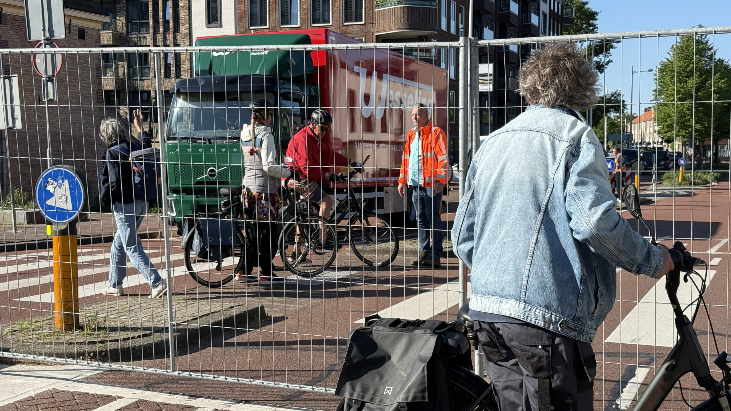 Mensen wachten met fietsen achter een hek bij een weg; op de achtergrond is een vrachtwagen te zien met een fel oranje jas drager ernaast en een verkeersbord met een looprichting.