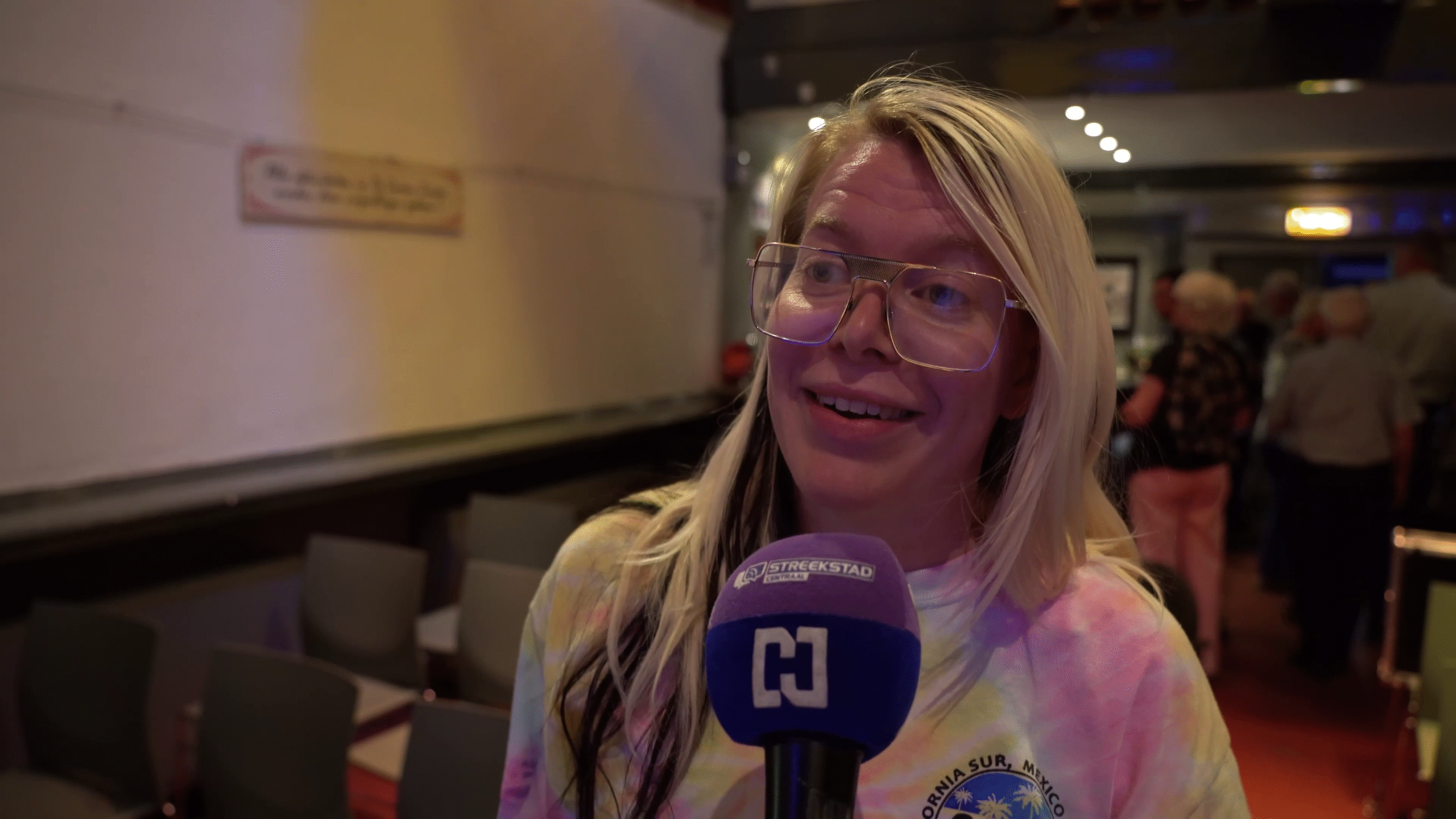 Een vrouw met lang blond haar en een bril spreekt in een microfoon met het logo van "Streekstad" erop, in een zaal met stoelen en mensen op de achtergrond.