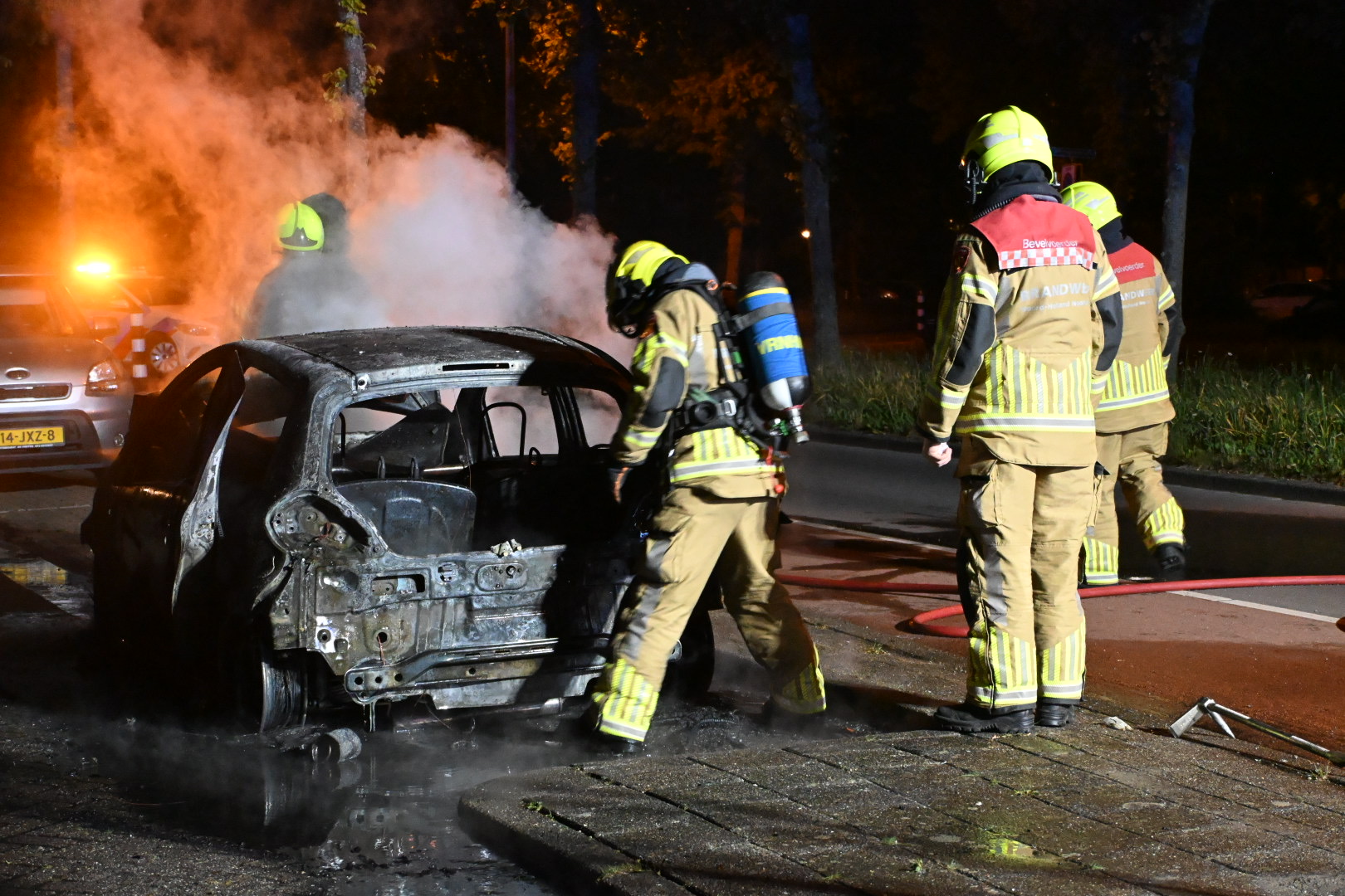 Brandweerlieden in gele pakken blussen een brandende auto, die zwaar beschadigd is, terwijl rook opstijgt en een politieauto met knipperende lichten op de achtergrond staat geparkeerd.