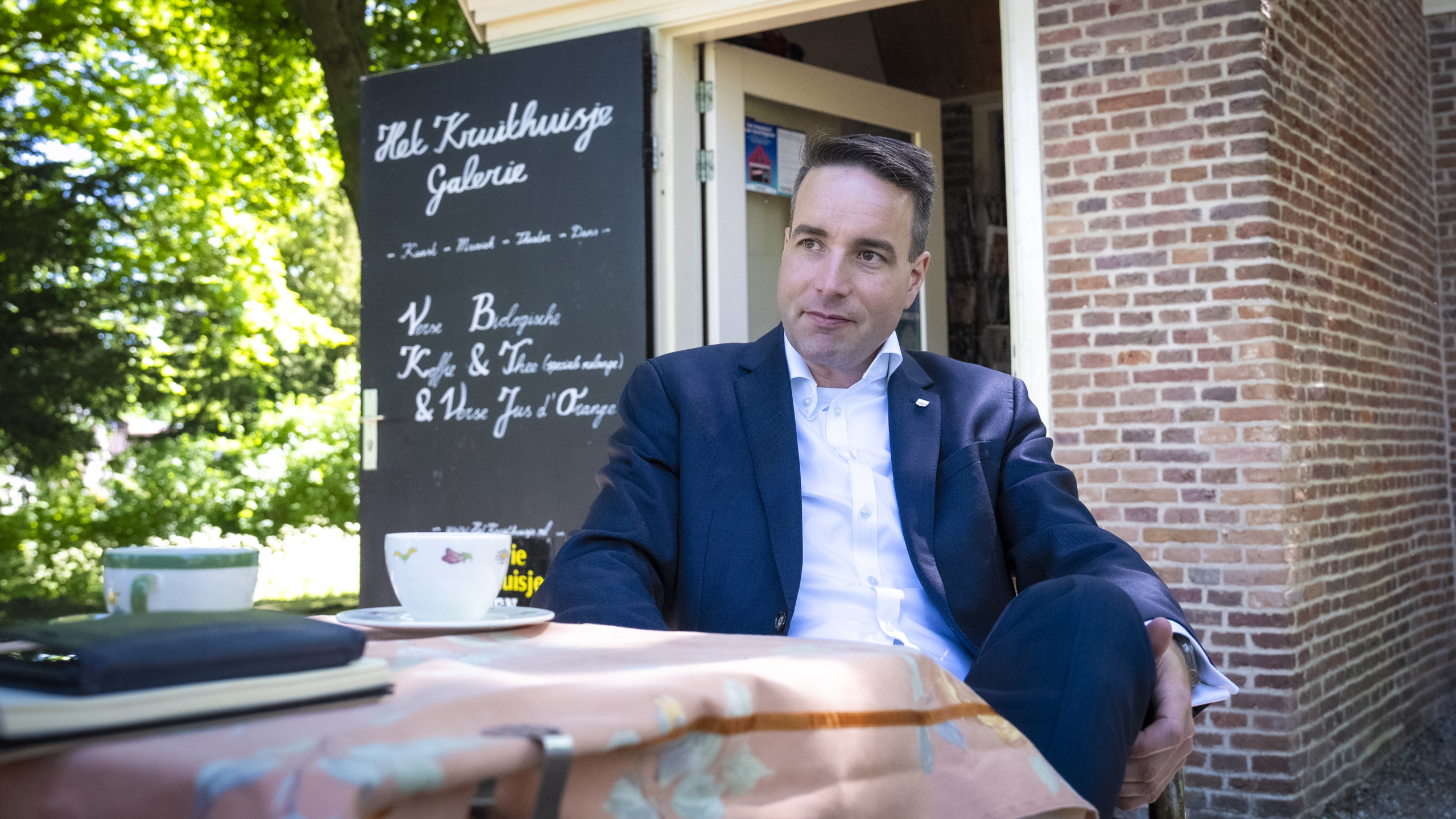 Een man in een donker pak zit ontspannen aan een tafel buiten, bij een gebouw met een bord waarop "Het Kruidhuysje Galerie" staat, omgeven door groene bomen.
