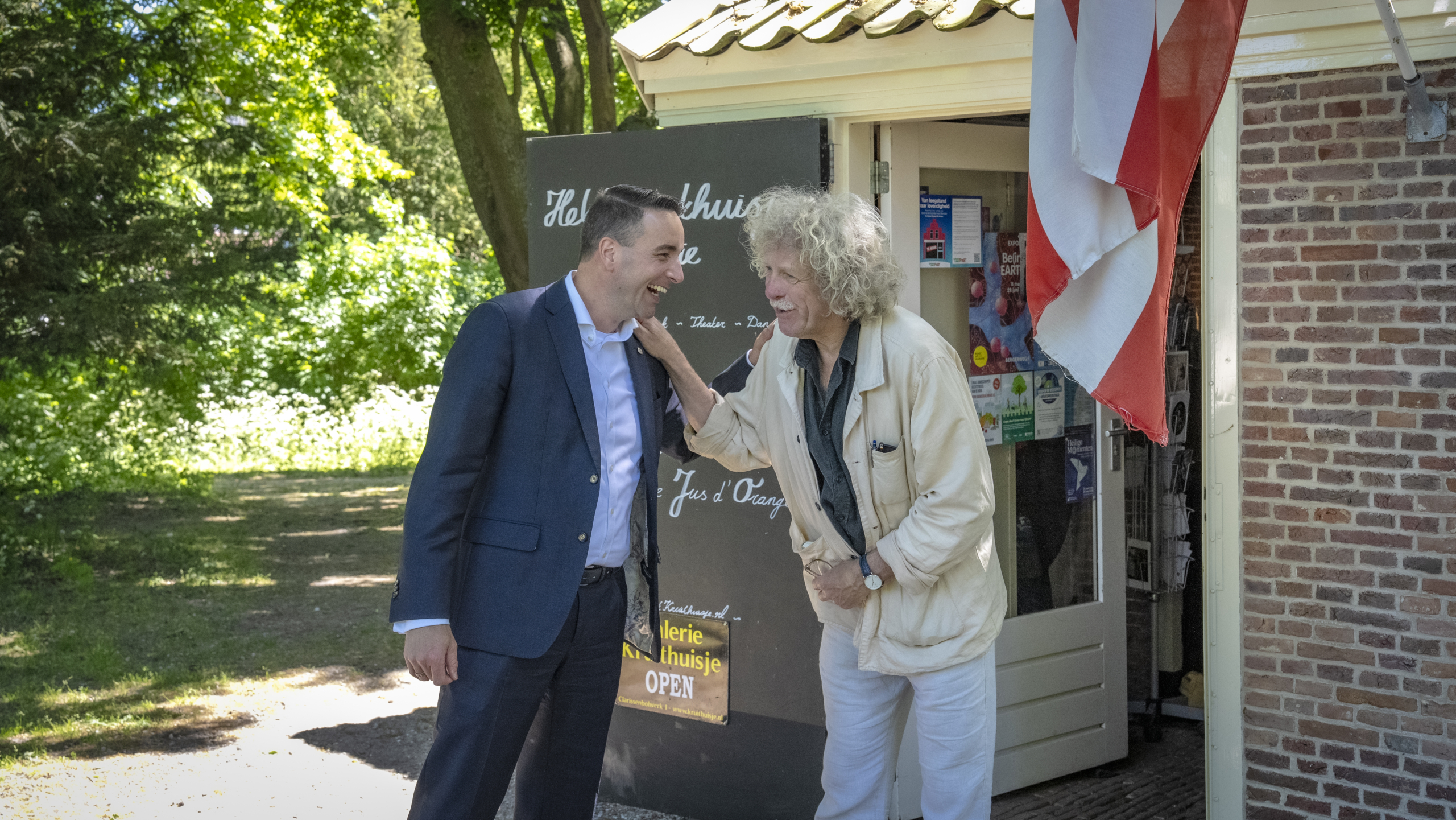 Twee mannen lachen en praten buiten een historisch gebouw met een Nederlandse vlag en een bord met de tekst "Galerie Kleinste Huisje OPEN".