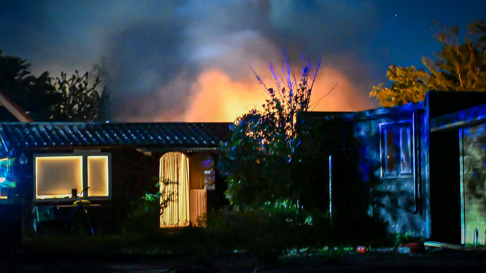 Nachtelijk tafereel met een brand waarbij vlammen achter een huis zichtbaar zijn, omringd door bomen en rook.