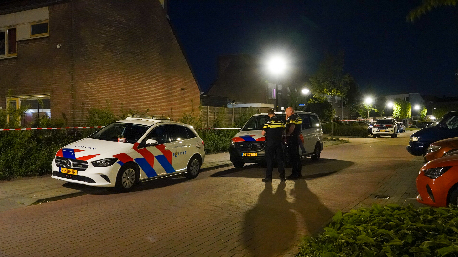 Twee politieauto's staan geparkeerd langs de straat in een woonwijk, met twee politieagenten die in gesprek zijn onder een straatlantaarn bij nachtelijke omstandigheden.