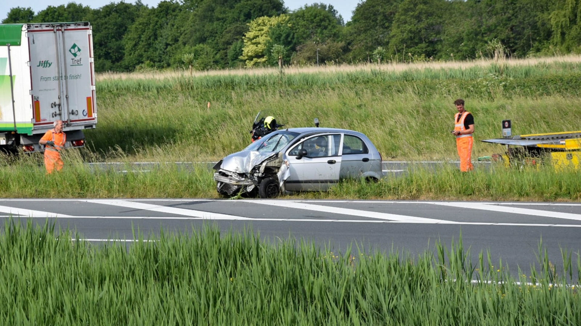 Een beschadigde zilverkleurige auto staat naast de snelweg, omgeven door gras en struiken. Er zijn drie personen in veiligheidskleding in de buurt, en er is ook een vrachtwagen en een sleepwagen aanwezig. Op de achtergrond is een groene, bosrijke omgeving te zien.