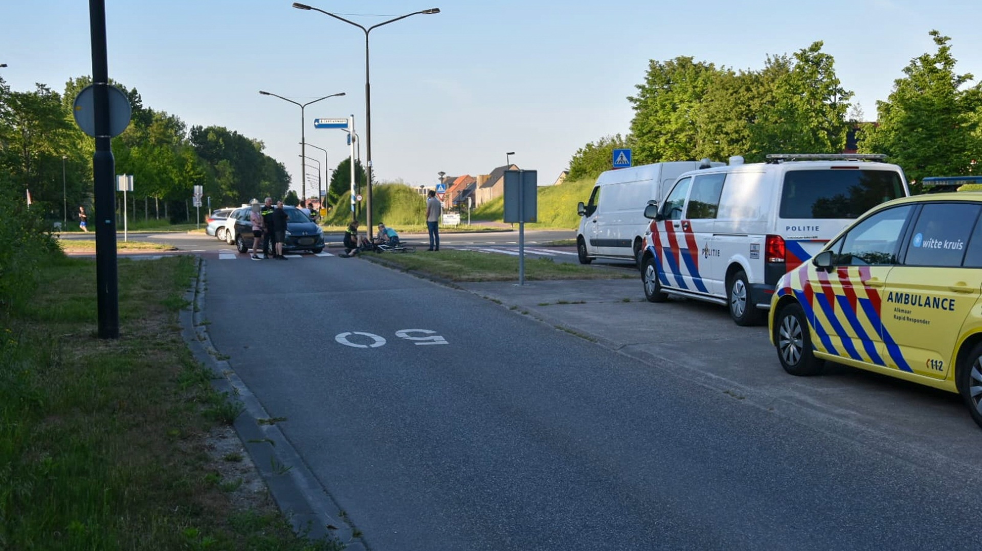 Een ongeval op een weg met een auto en fietsers, waarbij een politie- en ambulancevoertuig aanwezig zijn. Mensen staan rond de auto terwijl een slachtoffer op de grond zit. Er zijn bomen en verkeersborden zichtbaar in de omgeving.