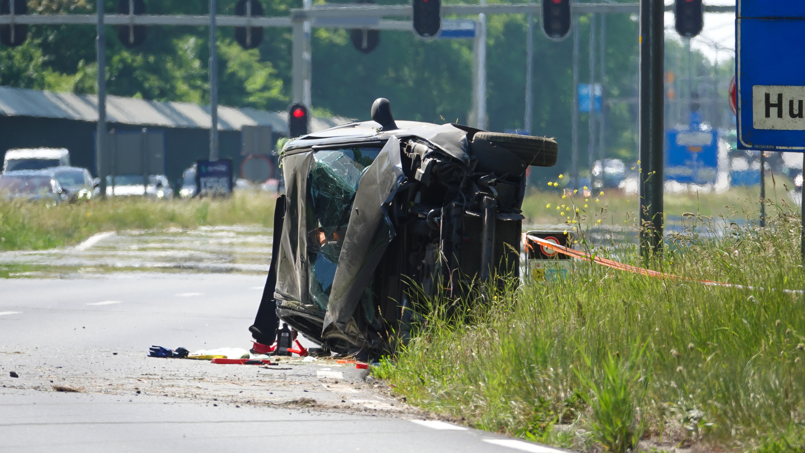 Een auto ligt op zijn kant op een weg met gras langs de berm. De voorruit is gebarsten en het voertuig lijkt zwaar beschadigd. Er zijn verkeerslichten en een verkeersbord zichtbaar op de achtergrond.