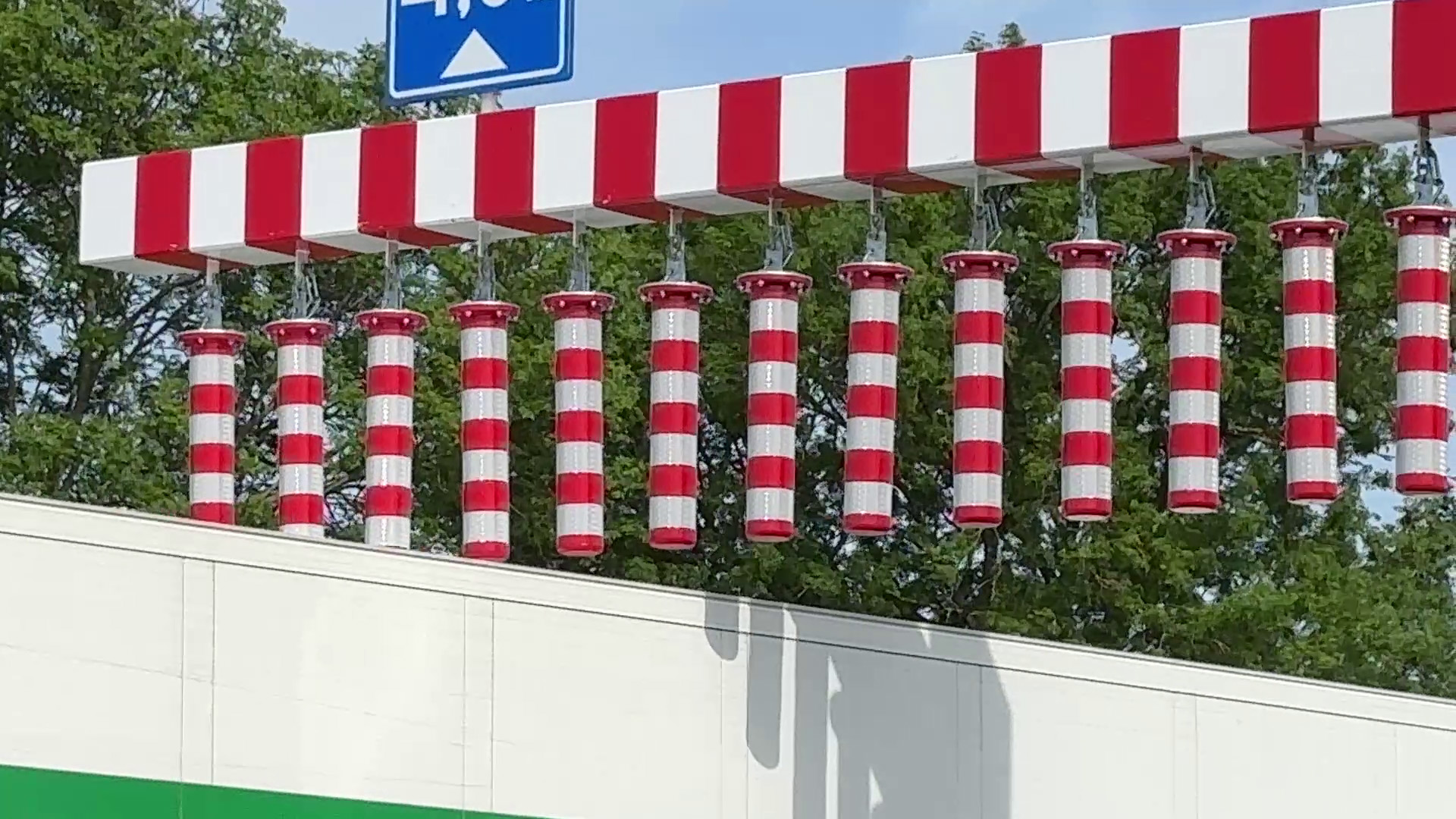 Wit-rood gestreepte waarschuwingspalen hangen boven een gebouw, met bomen en een blauw verkeersbord op de achtergrond.