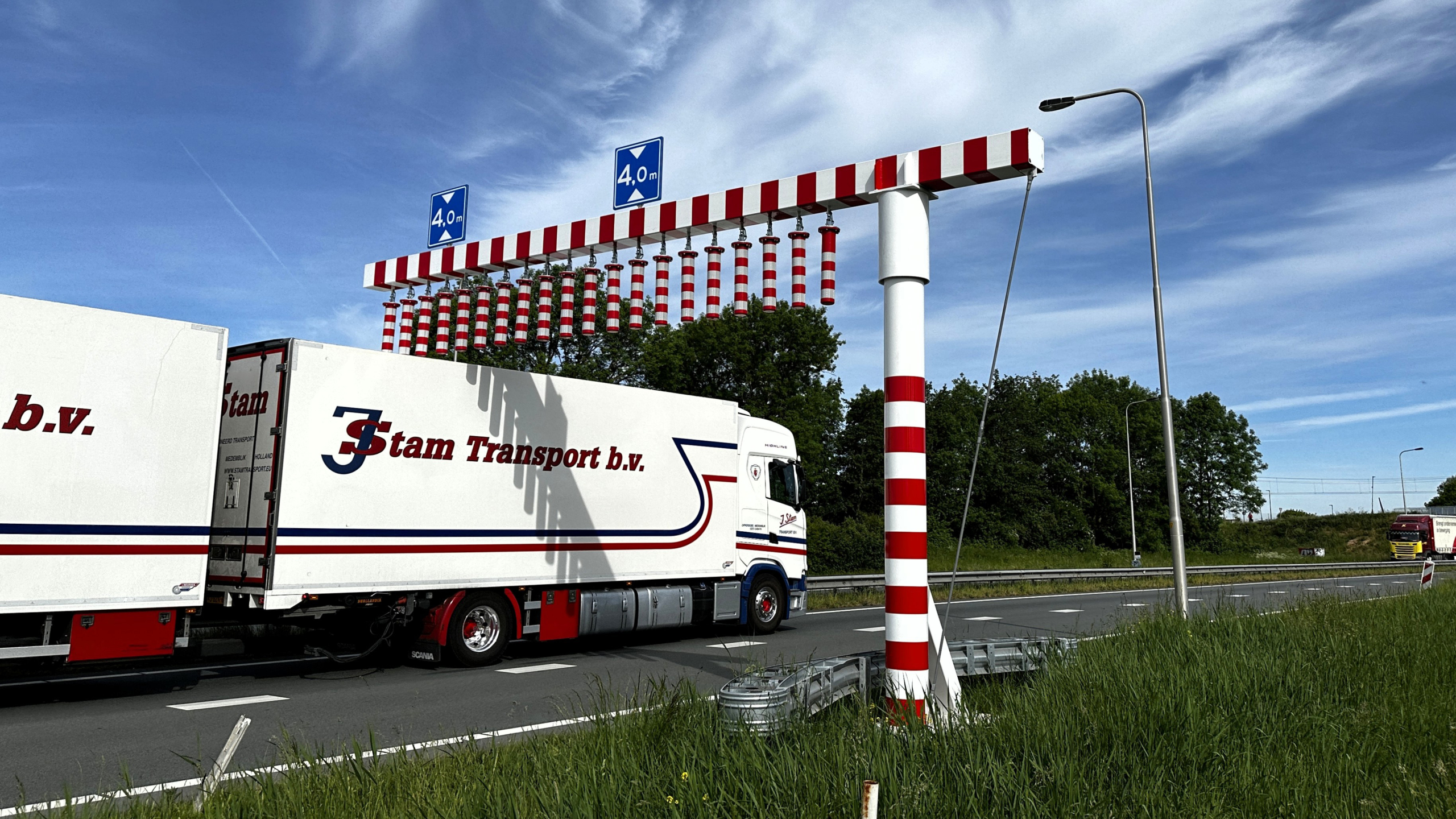 Een vrachtwagen passeert een hoogtebegrenzer van 4,0 meter hoogte met rood-witte strepen boven een snelweg, onder een blauwe lucht.
