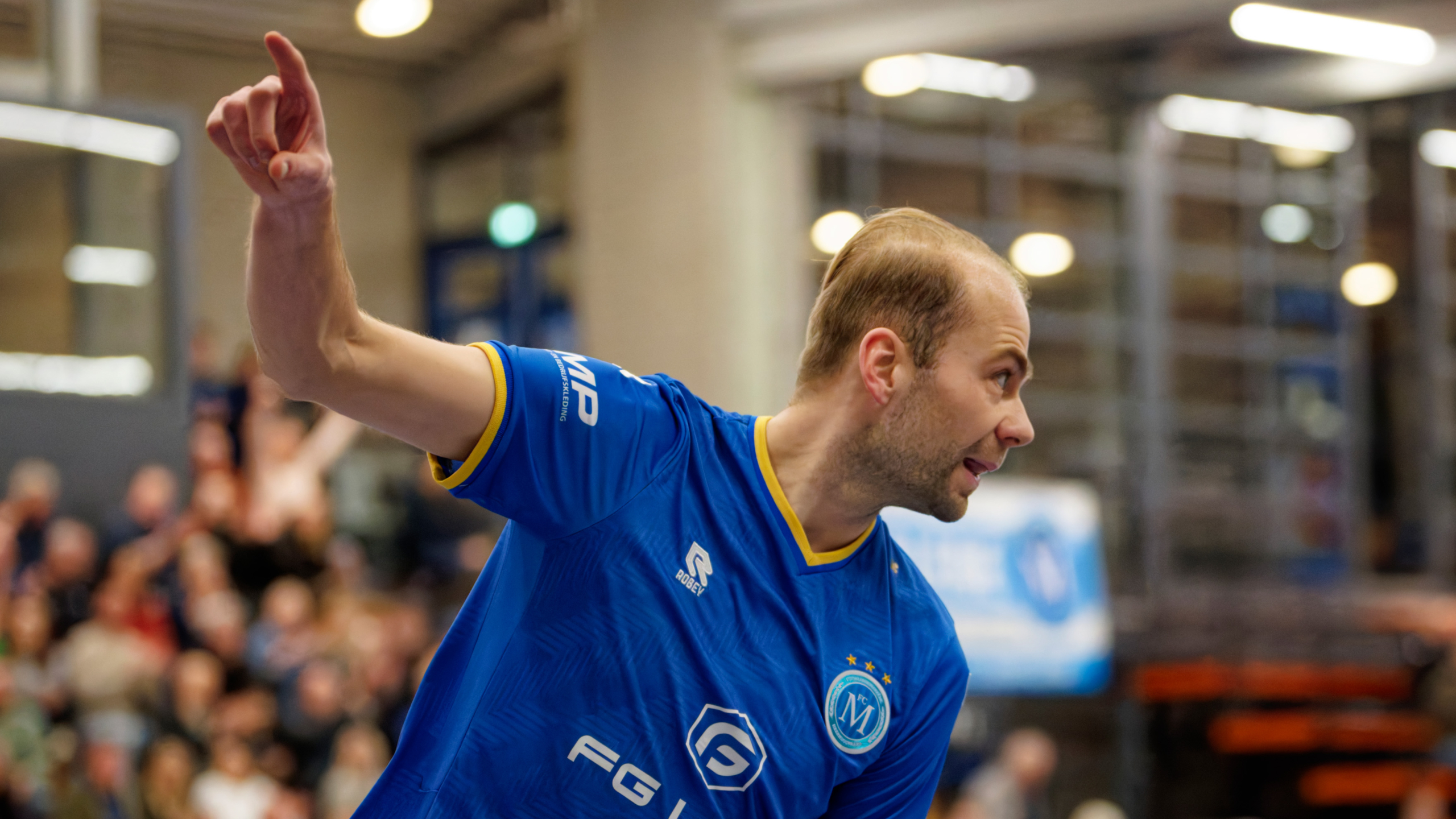Een man in een blauwe sportoutfit wijst energiek met zijn vinger omhoog in een sporthal met toeschouwers op de achtergrond.