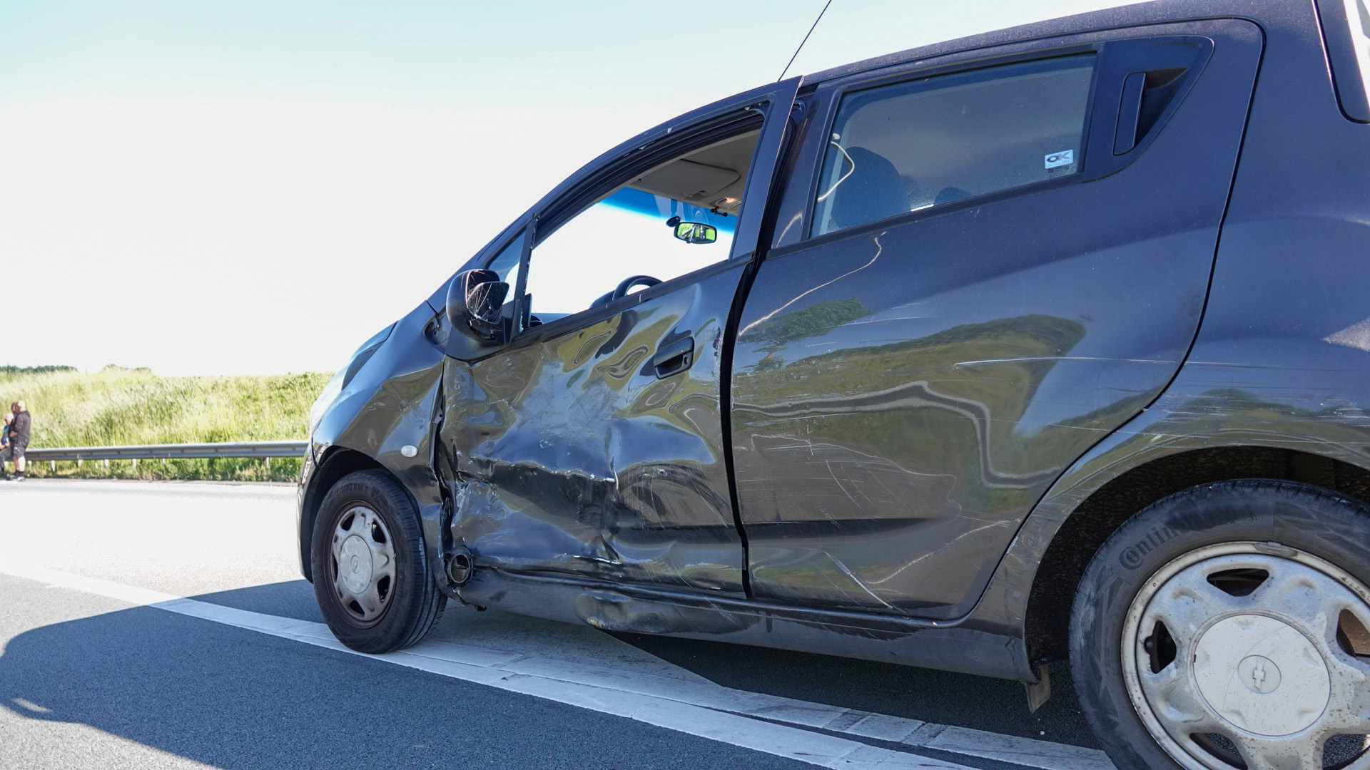 Een grijze auto met ernstige schade aan de zijkant staat stil naast de snelweg, met in de achtergrond een koppel dat langs de weg staat.