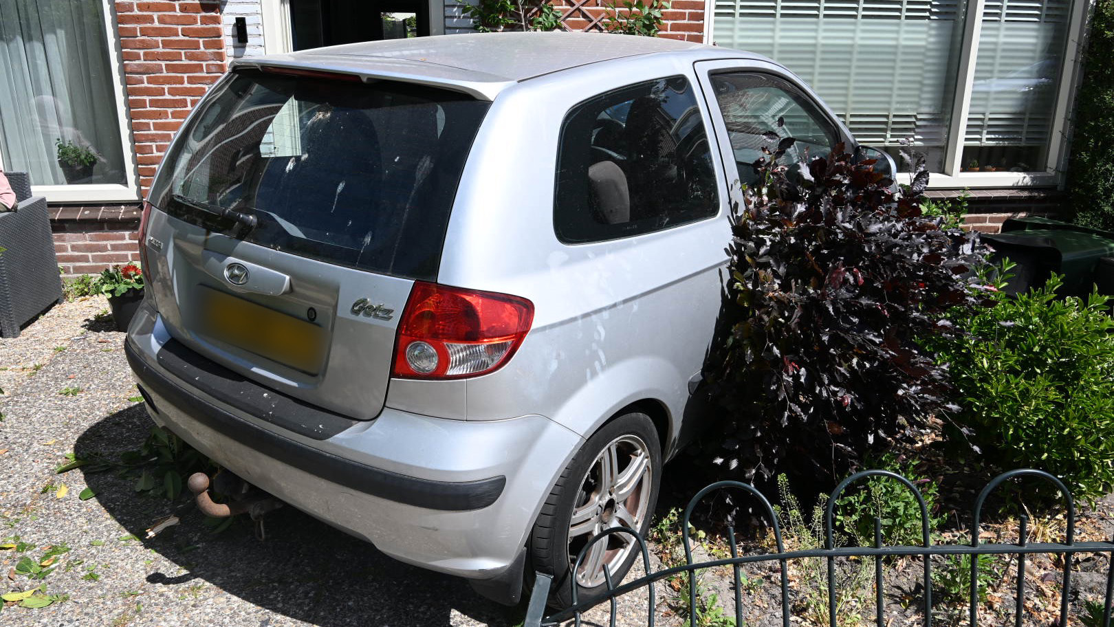Een zilveren auto van het merk Hyundai is tegen een struik en dicht bij een huis geparkeerd, met de achterzijde richting een verhard pad en aan de rechterkant een metalen hek.