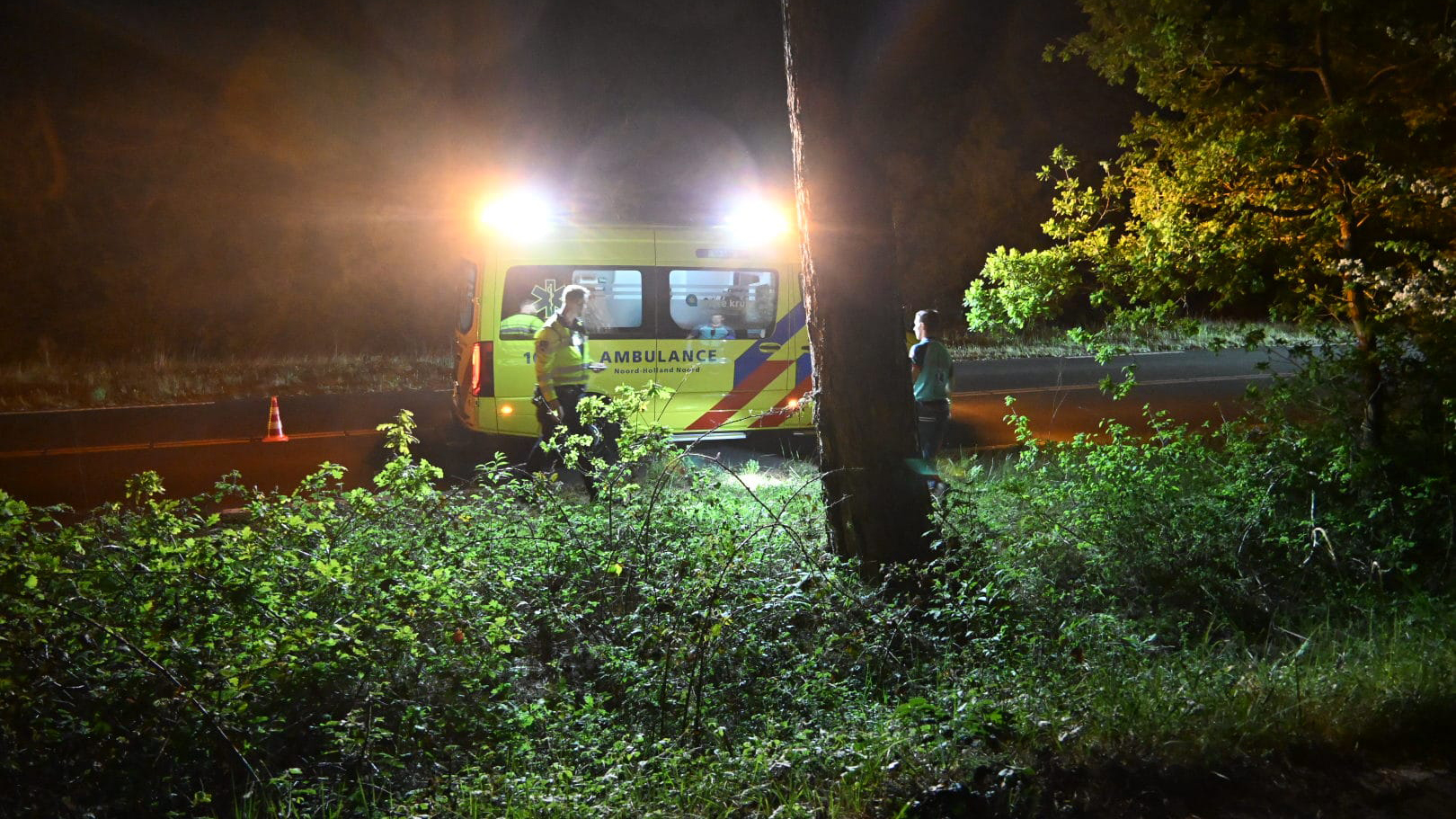 Nachtelijke scène met een ambulance langs de weg, verlicht met felle koplampen, terwijl een persoon in uniform naast het voertuig staat. De omgeving is omringd door bosjes en bomen, met een rustig wegdek.