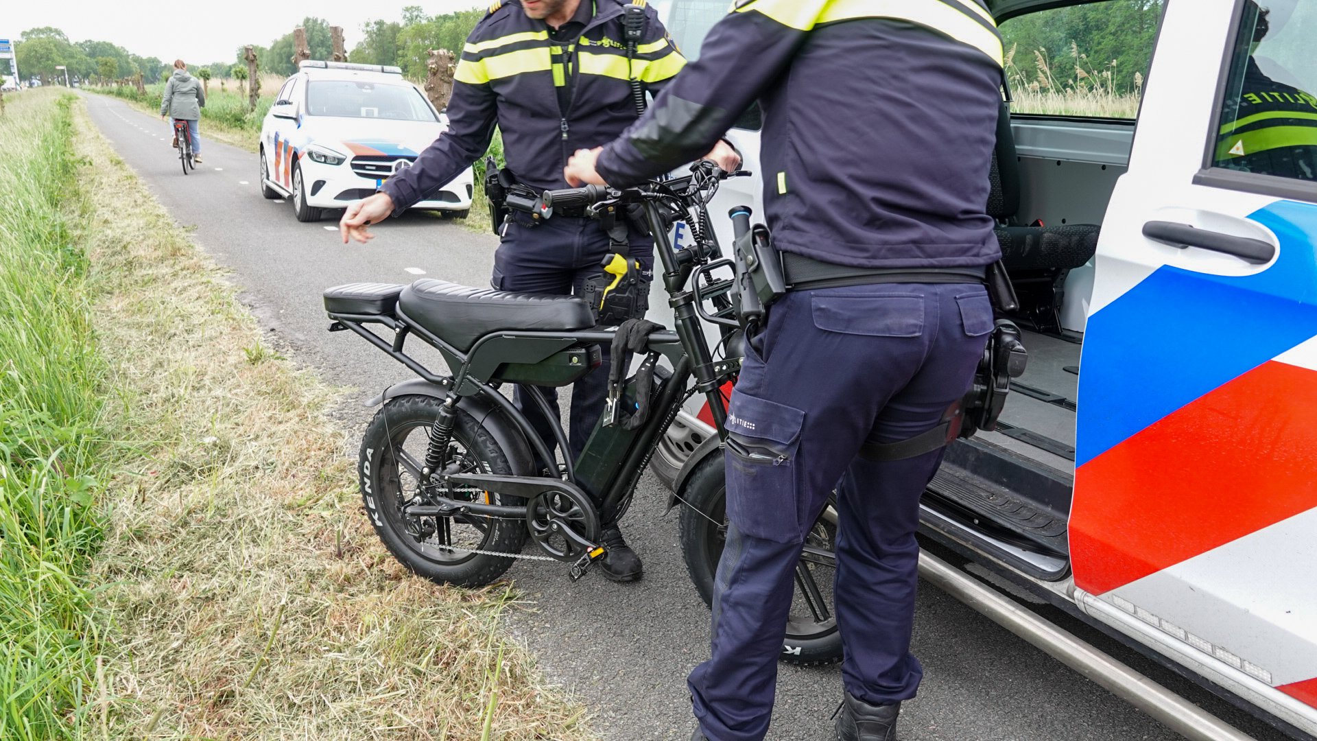 Twee politieagenten onderzoeken een elektrische fiets naast een politieauto op een landweg, terwijl een fietser passeert.