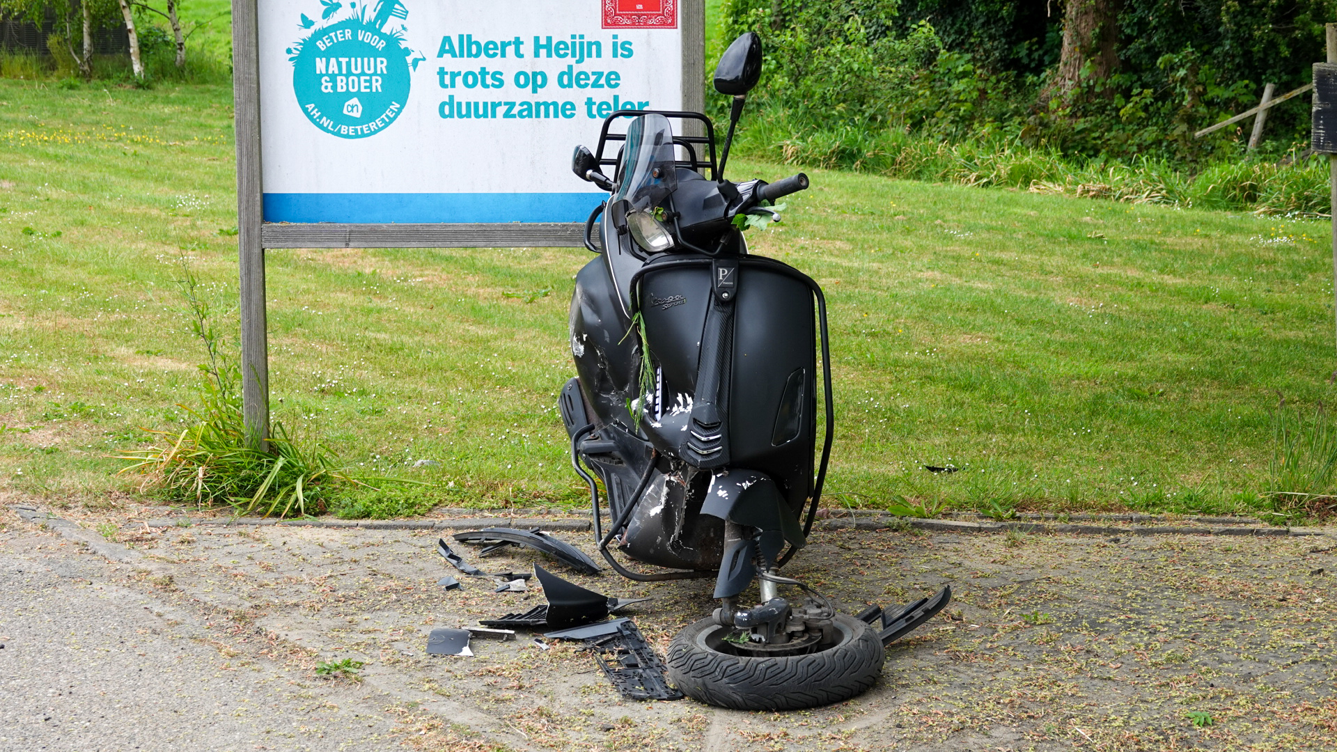 Beschadigde zwarte scooter met afgebroken onderdelen voor een bord met Albert Heijn-reclame, op een grasrijke locatie.