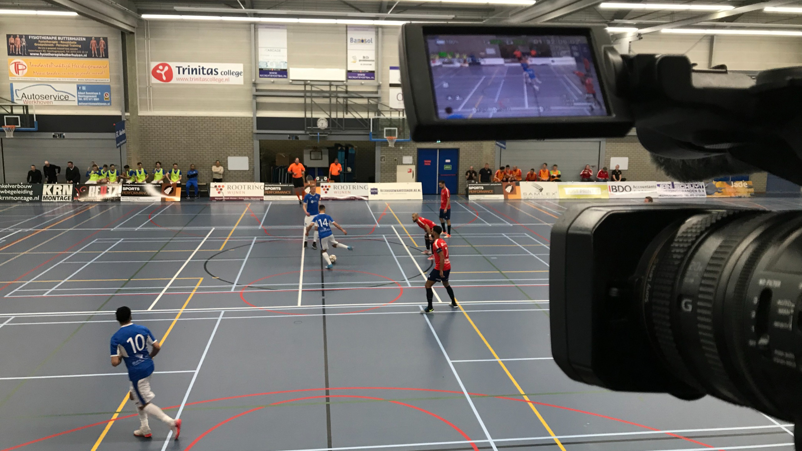 Een binnensportzaal waar een futsalwedstrijd plaatsvindt tussen teams in blauwe en rode outfits, vastgelegd door een videocamera.