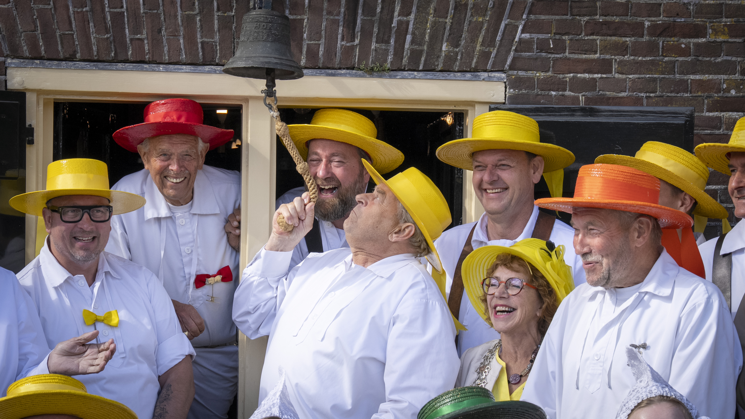 Een groep mensen in witte kleding en kleurrijke hoeden lacht terwijl een man aan een belring koord trekt voor een bakstenen gevel.